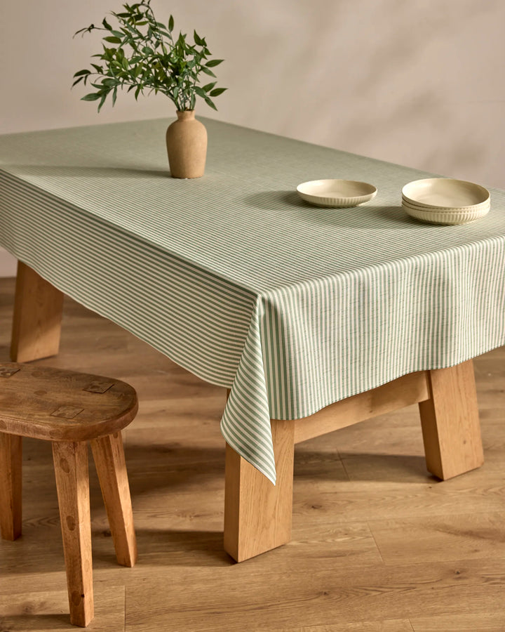 A wooden table draped in the Kailee Wright Mint Green Stripe eco-friendly tablecloth displays a beige vase with leafy branches and two white bowls, with a wooden stool beside it on a wooden floor.
