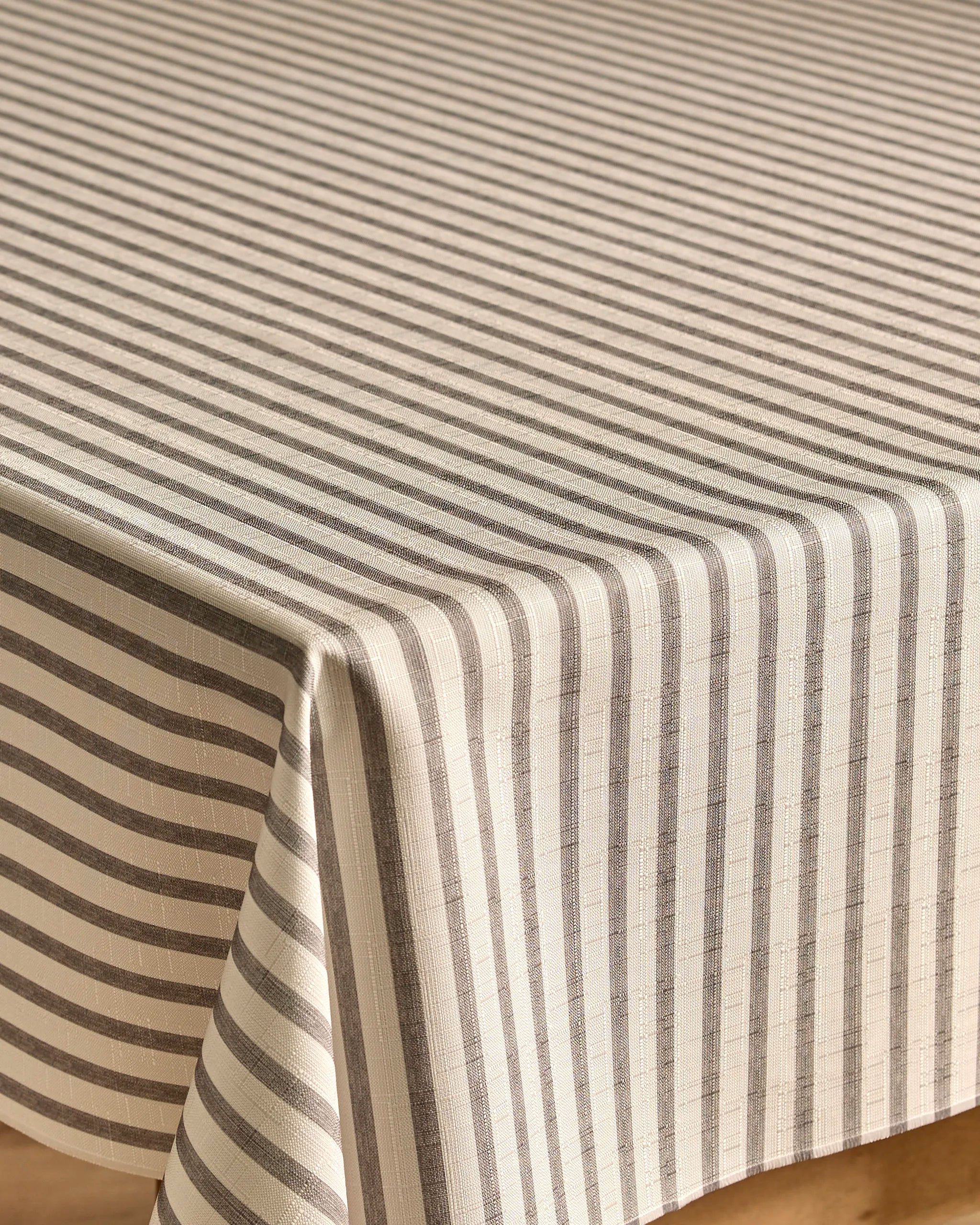 The Kailee Wright Stone Stripe tablecloth in beige features thin gray horizontal and vertical stripes, creating a neat geometric pattern. Lightweight and stain-resistant, it drapes elegantly over the table’s edge.