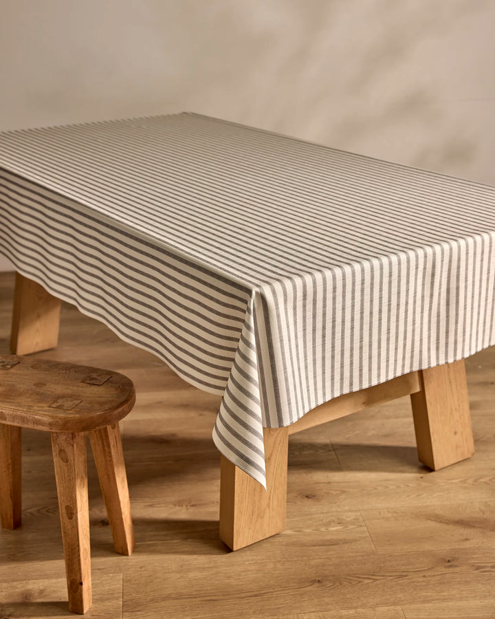 A Kailee Wright "Stone Stripe" gray and white striped tablecloth covers a wooden dining table beside a matching stool, all placed on a light wood floor with a neutral wall in the background.