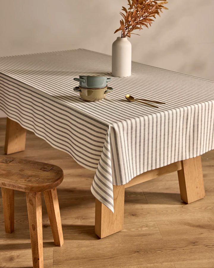 A Kailee Wright Stone Stripe gray and white stain-resistant tablecloth covers a wooden table, set with two stacked bowls, a plate, gold utensils, and a white vase with dried brown leaves. A wooden stool sits beside the table on the wooden floor.