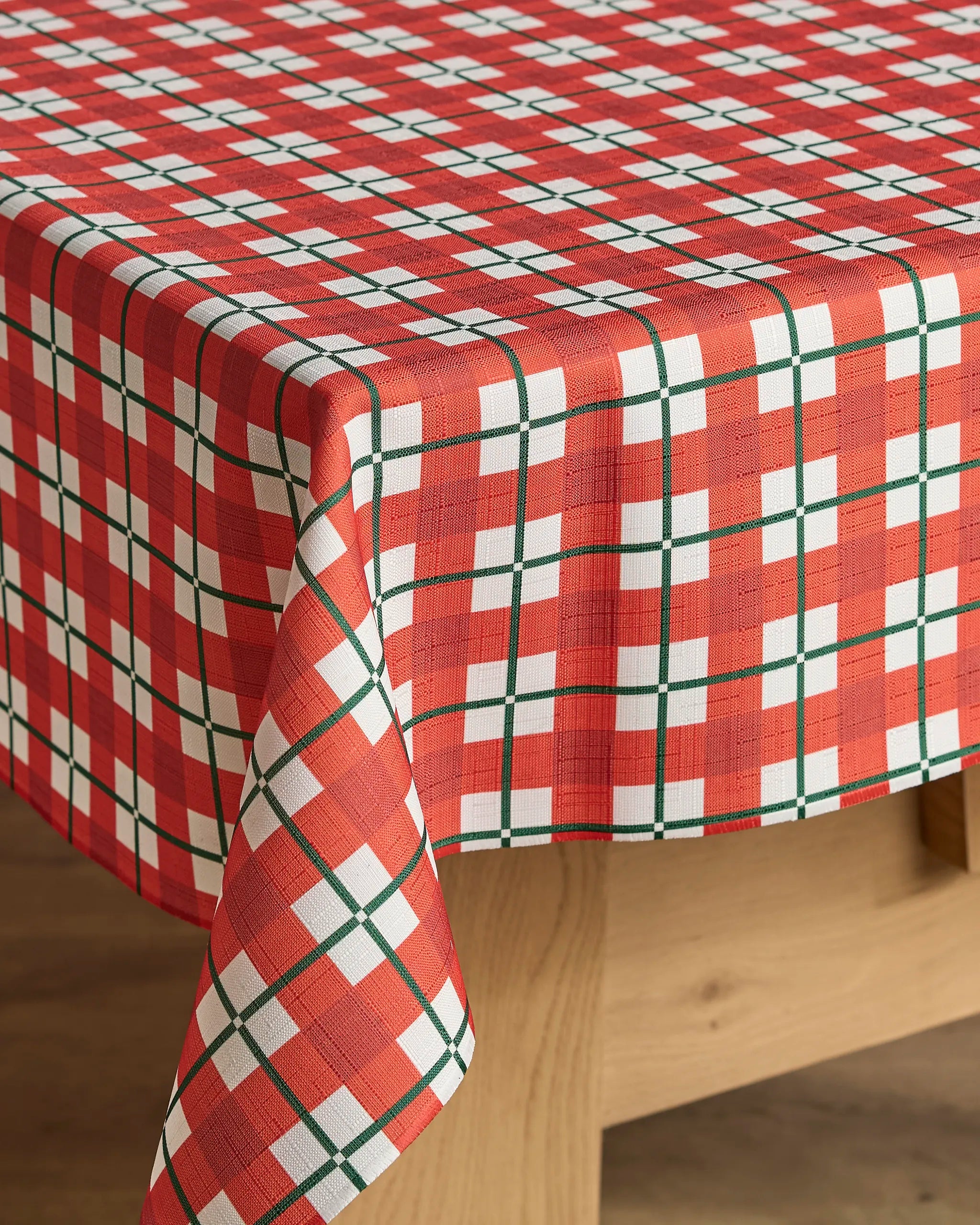 A wooden table topped with the Geometry Yuletide Plaid tablecloth, featuring a red, white, and green stain-resistant plaid design, drapes neatly over the edge onto a wooden floor.