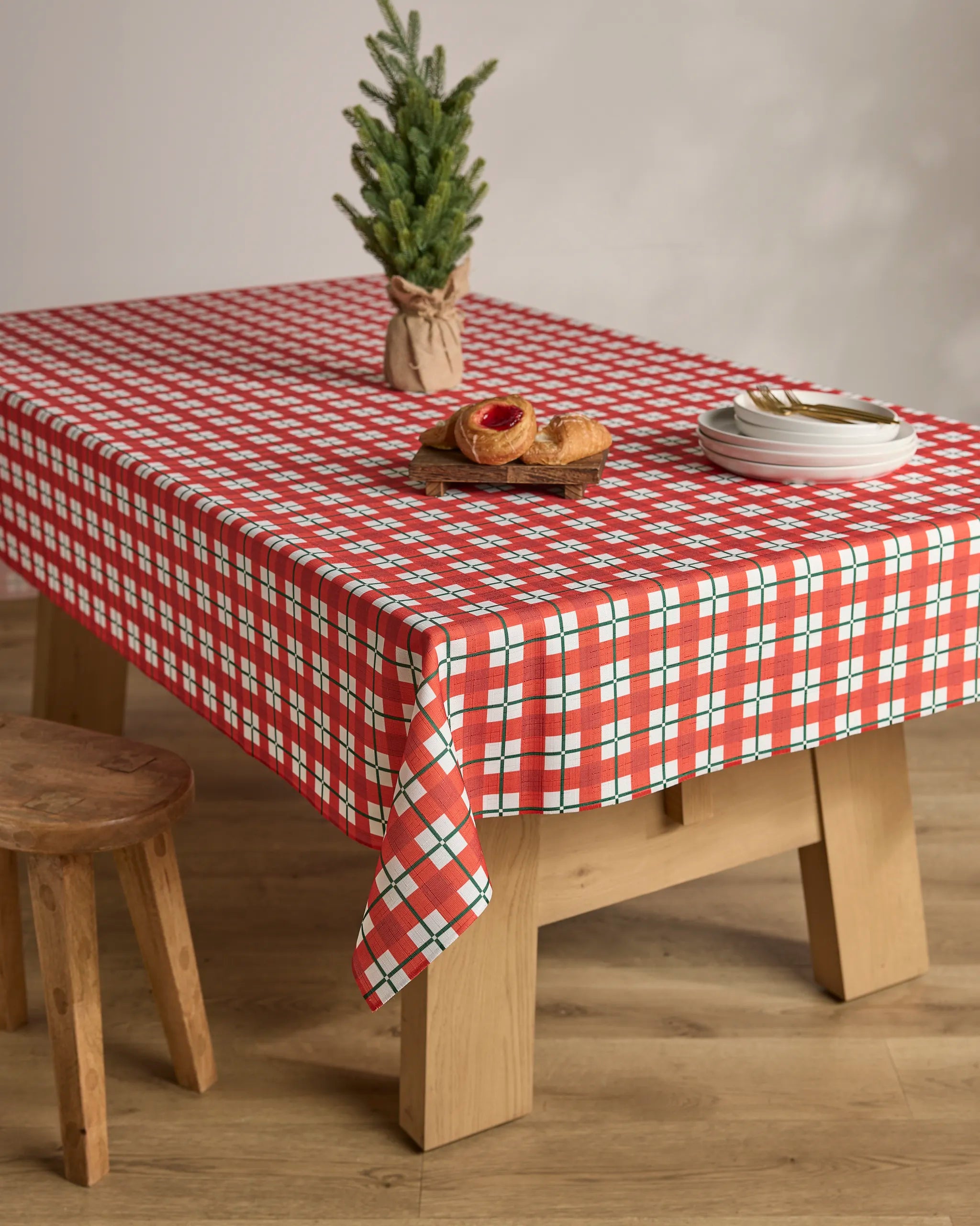 A wooden table with a Geometry Yuletide Plaid stain-resistant tablecloth, a small potted pine tree, pastries on a wooden tray, stacked white plates, and a wooden stool beside it on a wooden floor.
