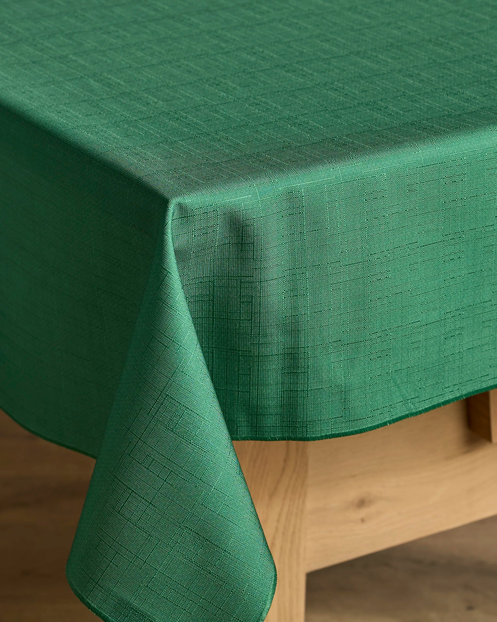 The Geometry Yuletide Spirit tablecloth, featuring a subtle textured pattern and stain-resistant green fabric, drapes neatly over a wooden table with one corner hanging down against a softly blurred background.