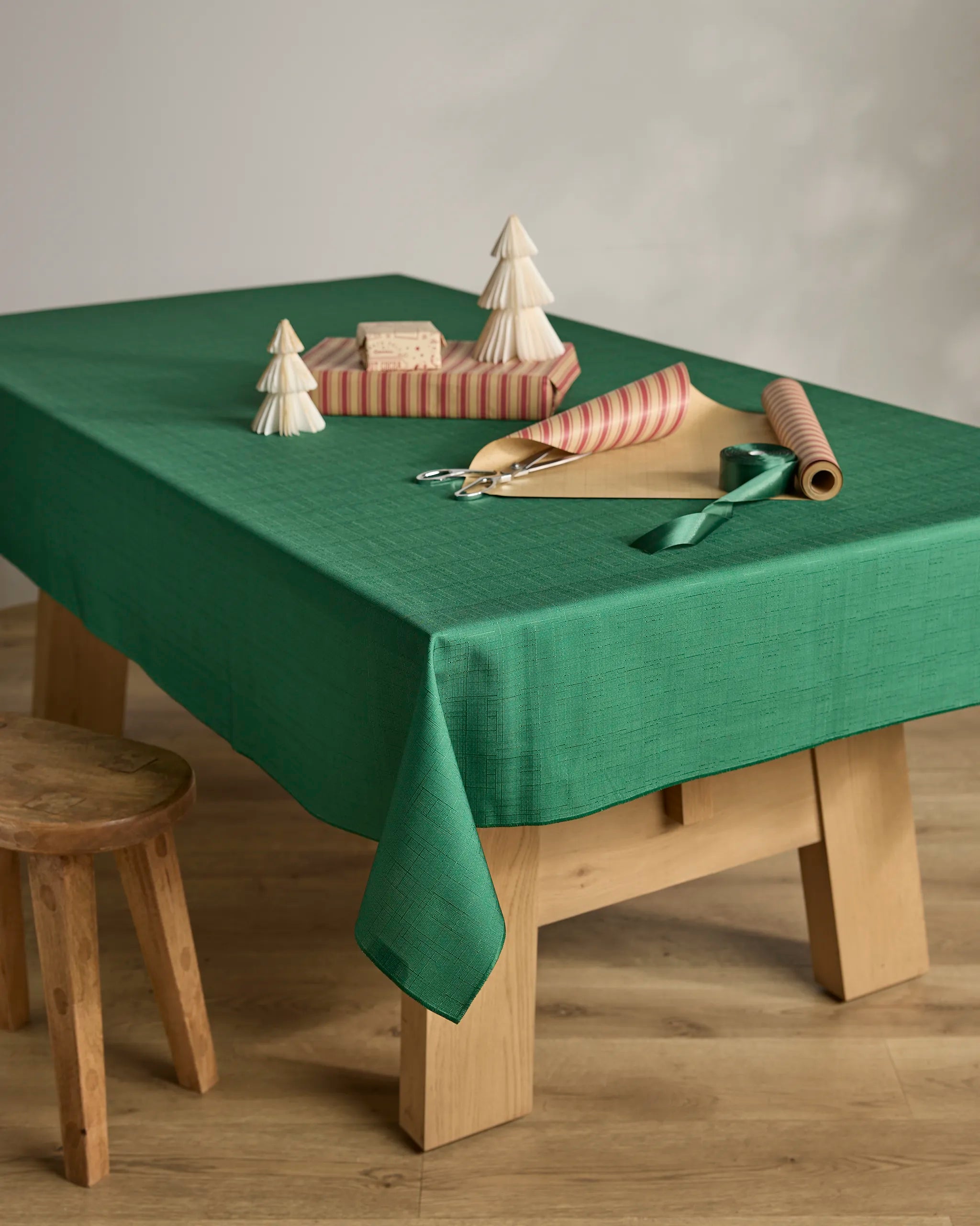 The Yuletide Spirit by Geometry features a stain-resistant green tablecloth on a wooden table, adorned with paper trees, pink-striped wrapped gifts, scissors, green ribbon, and paired with a small wooden stool on a light wood floor.