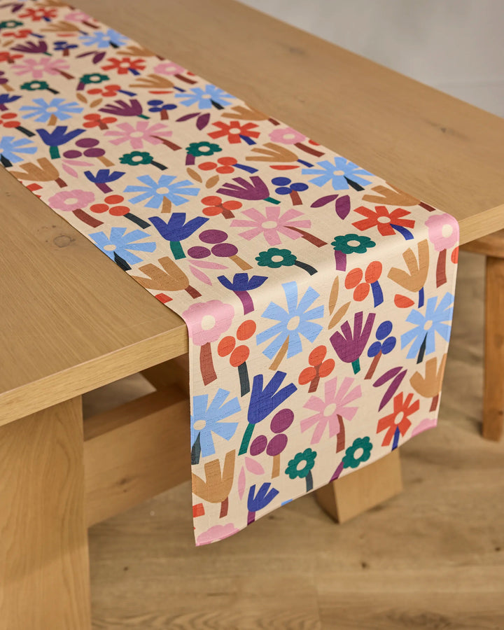 A wooden table with the Cool Flowers table runner by Jennifer Bouron, featuring a stain-resistant, light beige background with colorful stylized red, blue, purple, orange, and green flowers.