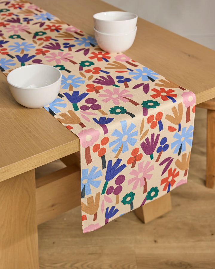 A light wooden table displays the Cool Flowers runner by Jennifer Bouron—stain-resistant, with bright blue, red, purple, and green blooms on beige—topped with three white bowls.