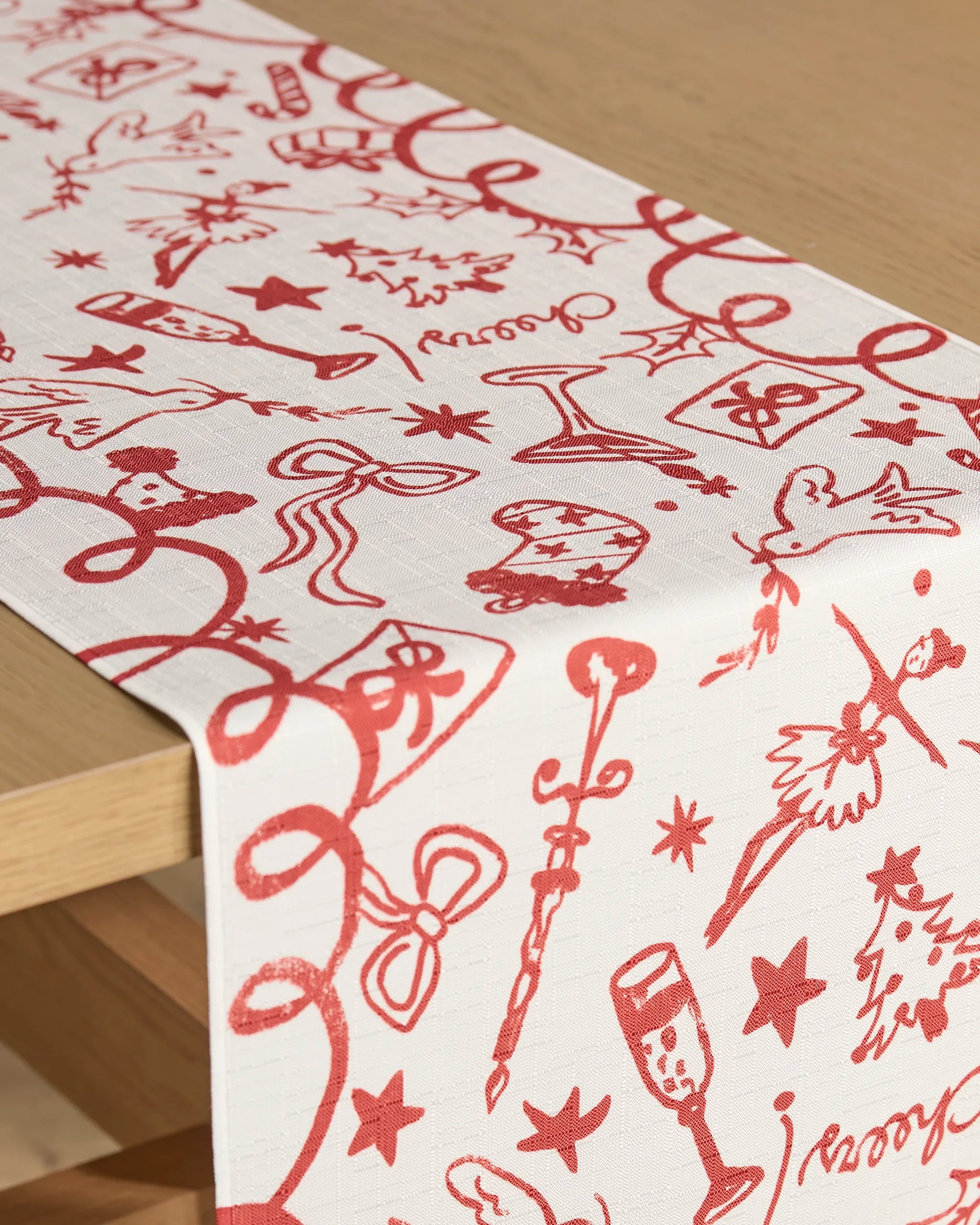The Geometry "Doodles for Holiday" stain-resistant table runner, featuring red festive illustrations, adds charm to a light wooden table.