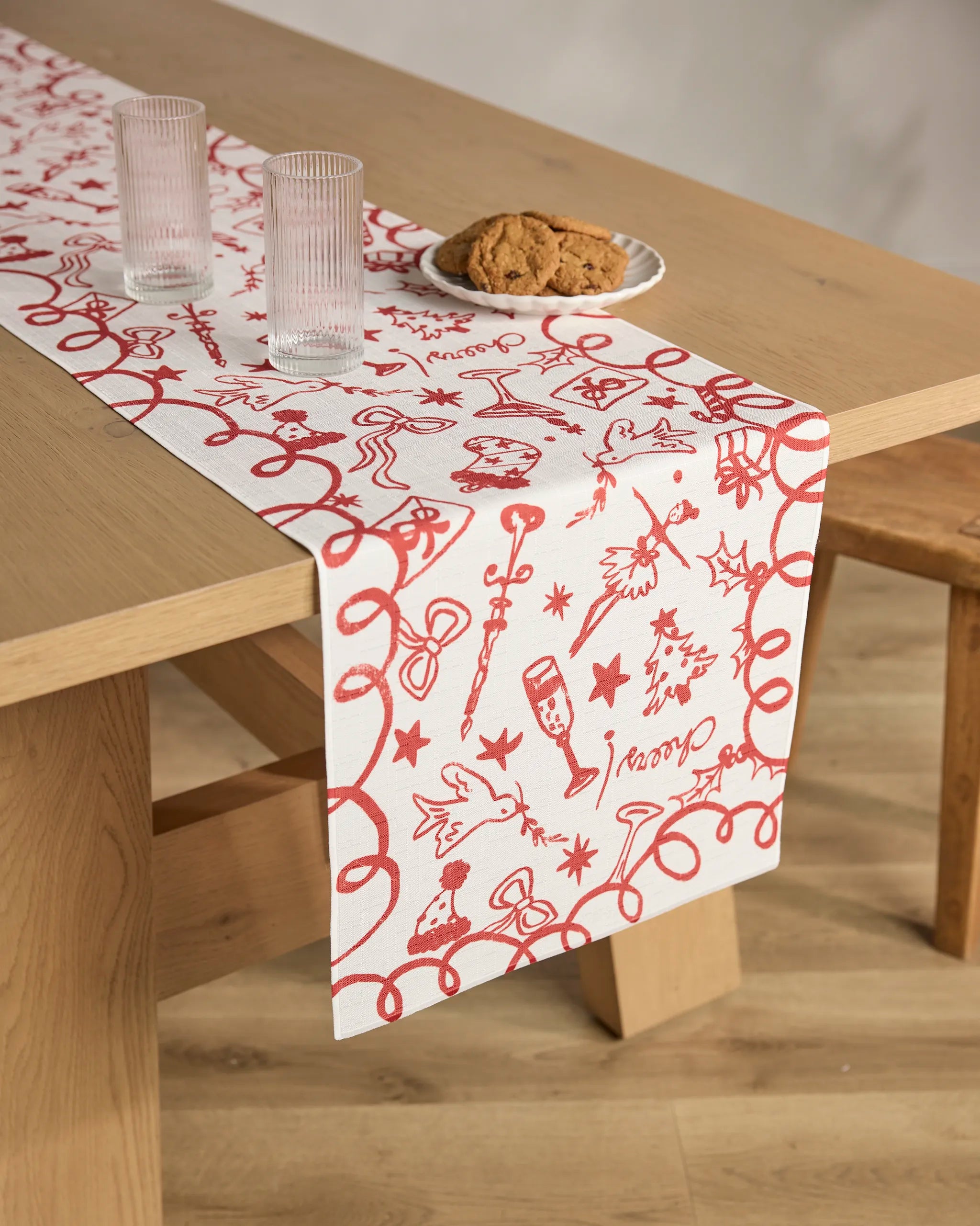 A wooden table with the Geometry "Doodles for Holiday" stain-resistant runner featuring red holiday designs, two clear water glasses, and cookies on a plate; a matching wooden bench sits nearby.