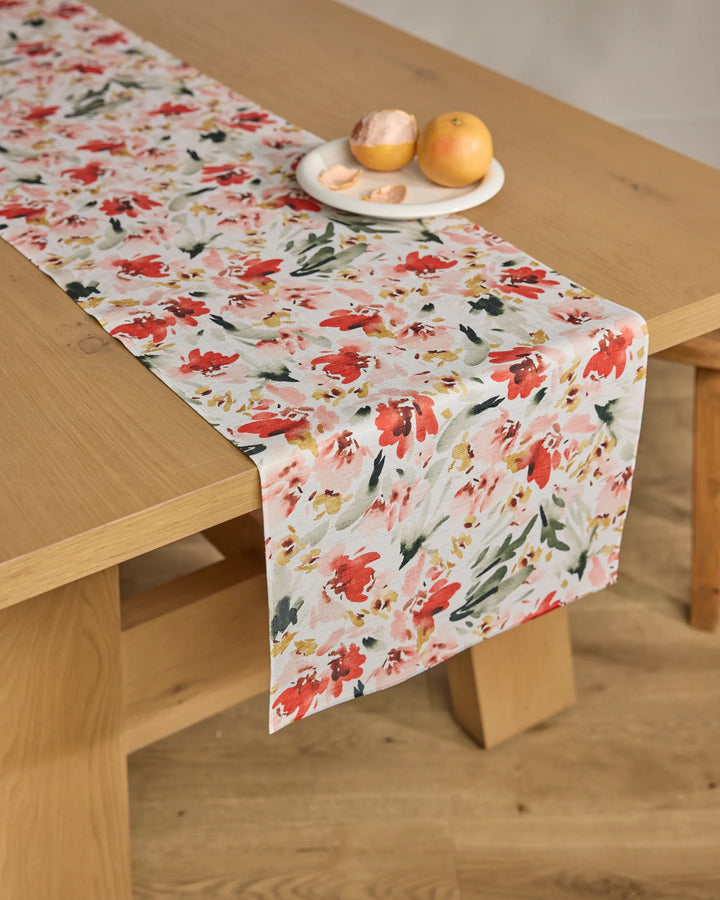 A wooden table displays the Melissa Johnson "Painted Garden" stain-resistant table runner with red, pink, and green floral watercolor. On it sits a white plate with a peach, an orange, and peach slices.