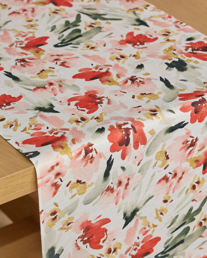 A close-up of the Melissa Johnson "Painted Garden" stain-resistant table runner, featuring a vibrant floral pattern in red, pink, green, and yellow, draped over a light wooden table.