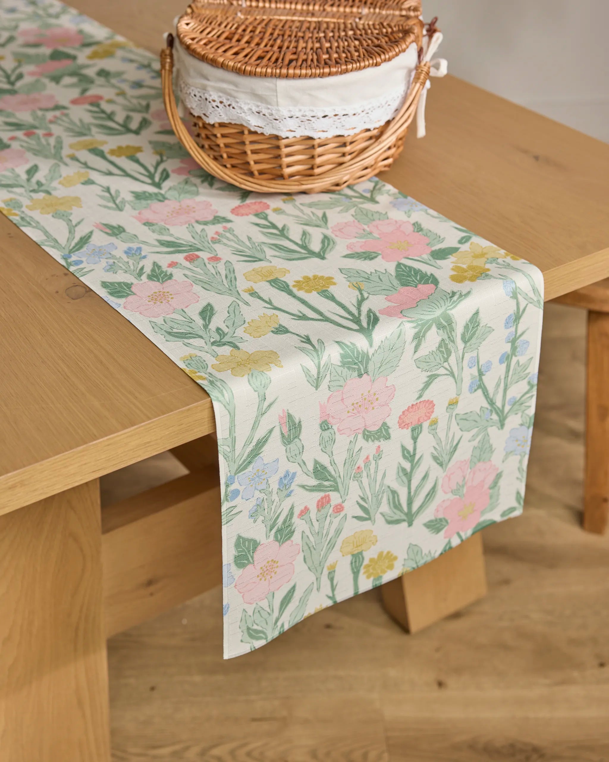 A wooden table features the "Wildflowers in Bloom" stain-resistant table runner by Jasmine Blooms Designs and a wicker basket with a lace-trimmed cloth, all set atop a light wood floor.