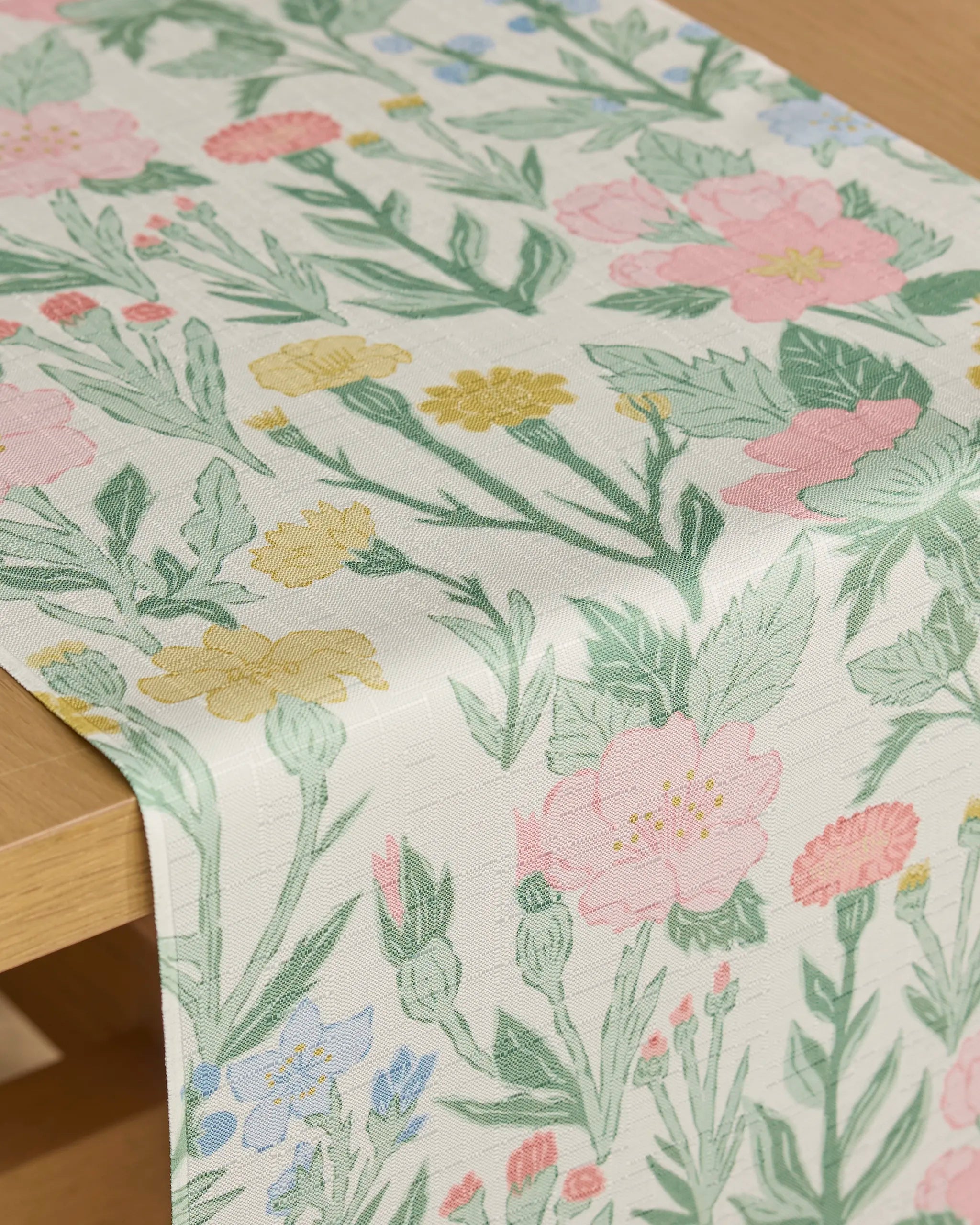 The "Wildflowers in Bloom" stain-resistant table runner by Jasmine Blooms Designs features pink, yellow, and blue floral prints with green leaves, beautifully shown on a light wooden table.