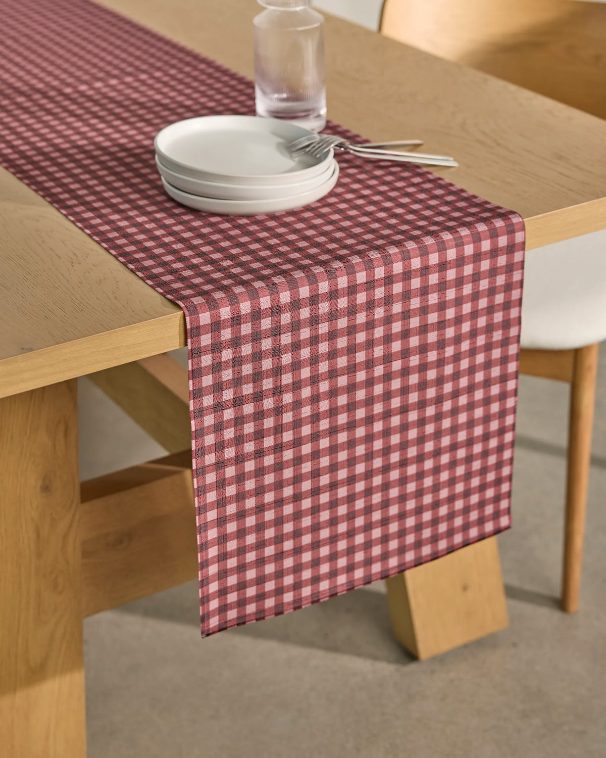 A wooden table is adorned with the Berry Check stain-resistant table runner from Geometry, featuring a red and white checkered pattern. On top are stacked white dishes, a drinking glass, a fork, and a spoon. A light-colored chair sits beside the table.