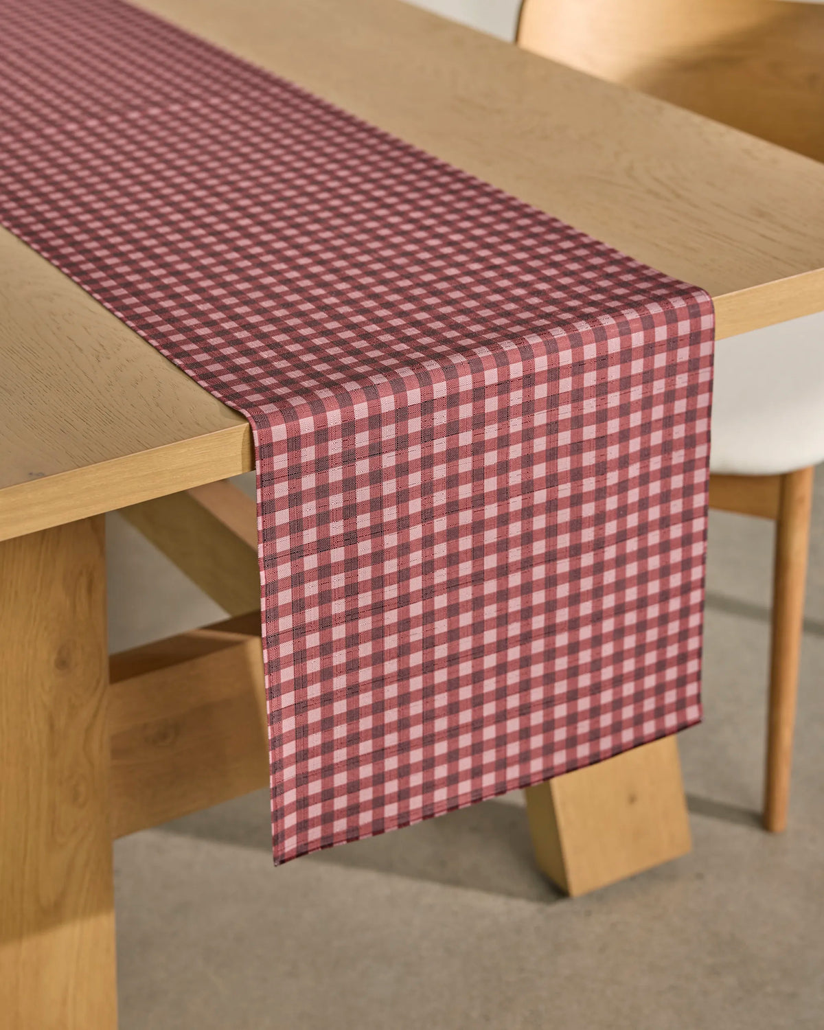 A light-colored chair sits next to a wooden table adorned with the Geometry Berry Check stain-resistant table runner, its red and white checkered pattern draped neatly over the edge.