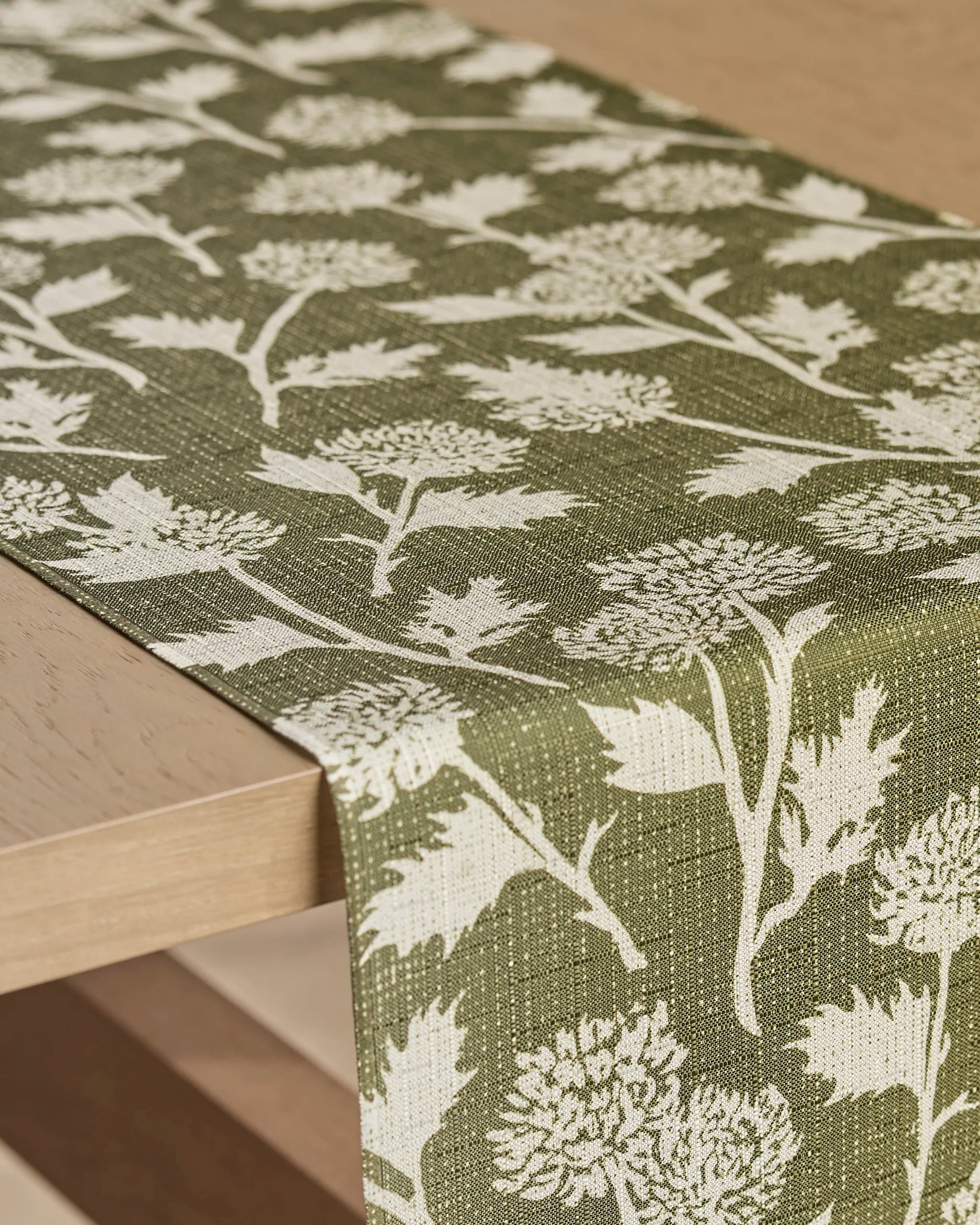 The Geometry Bloom Thistle Green table runner, stain-resistant and adorned with detailed white botanical flowers and leaves, is draped over a light wooden table.