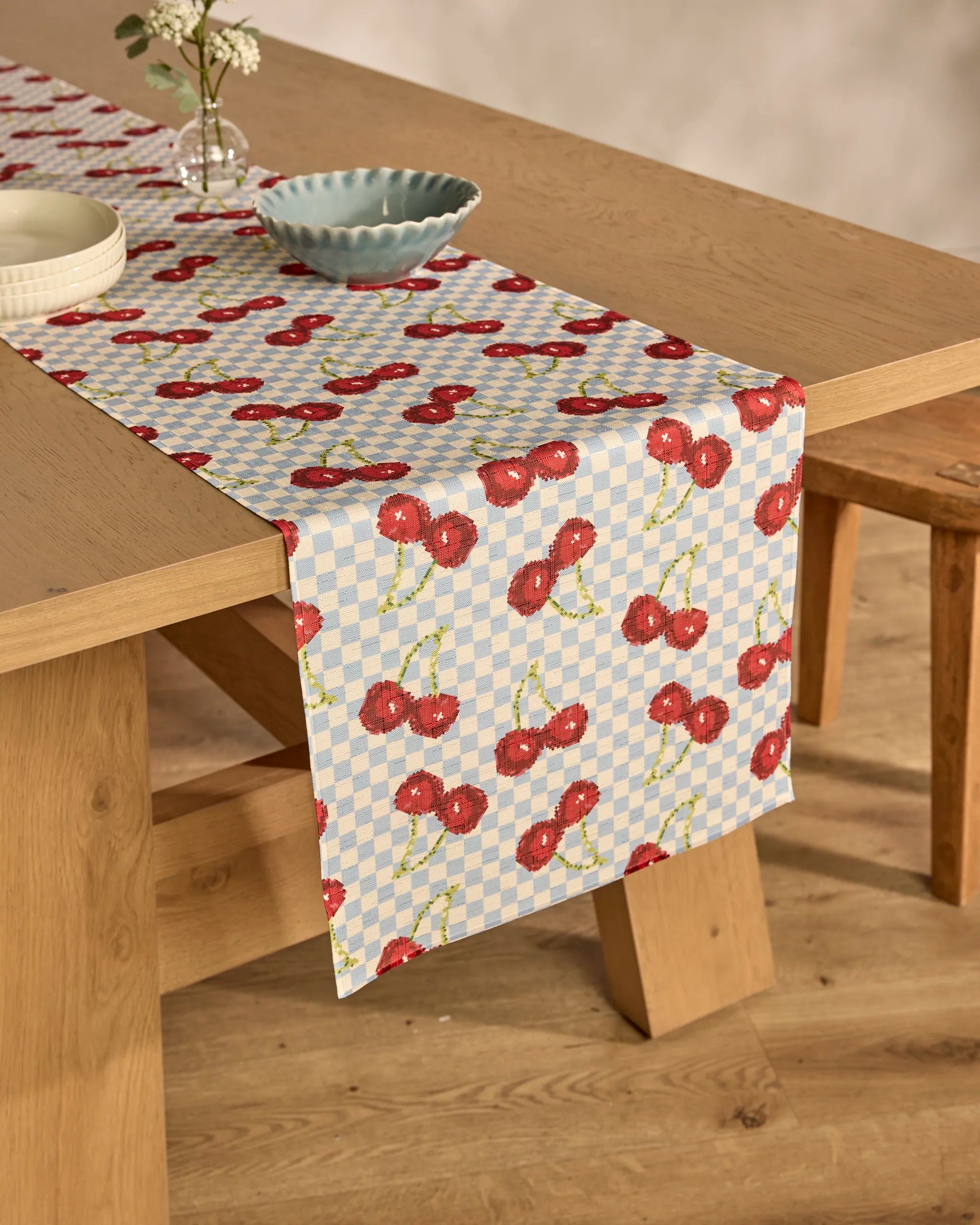 The Geometry "Cherry Cola Days" stain-resistant checkered table runner with red cherries decorates a wooden dining table, which is set with a small vase of flowers, a white plate, a blue scalloped bowl, and accompanied by a wooden bench.