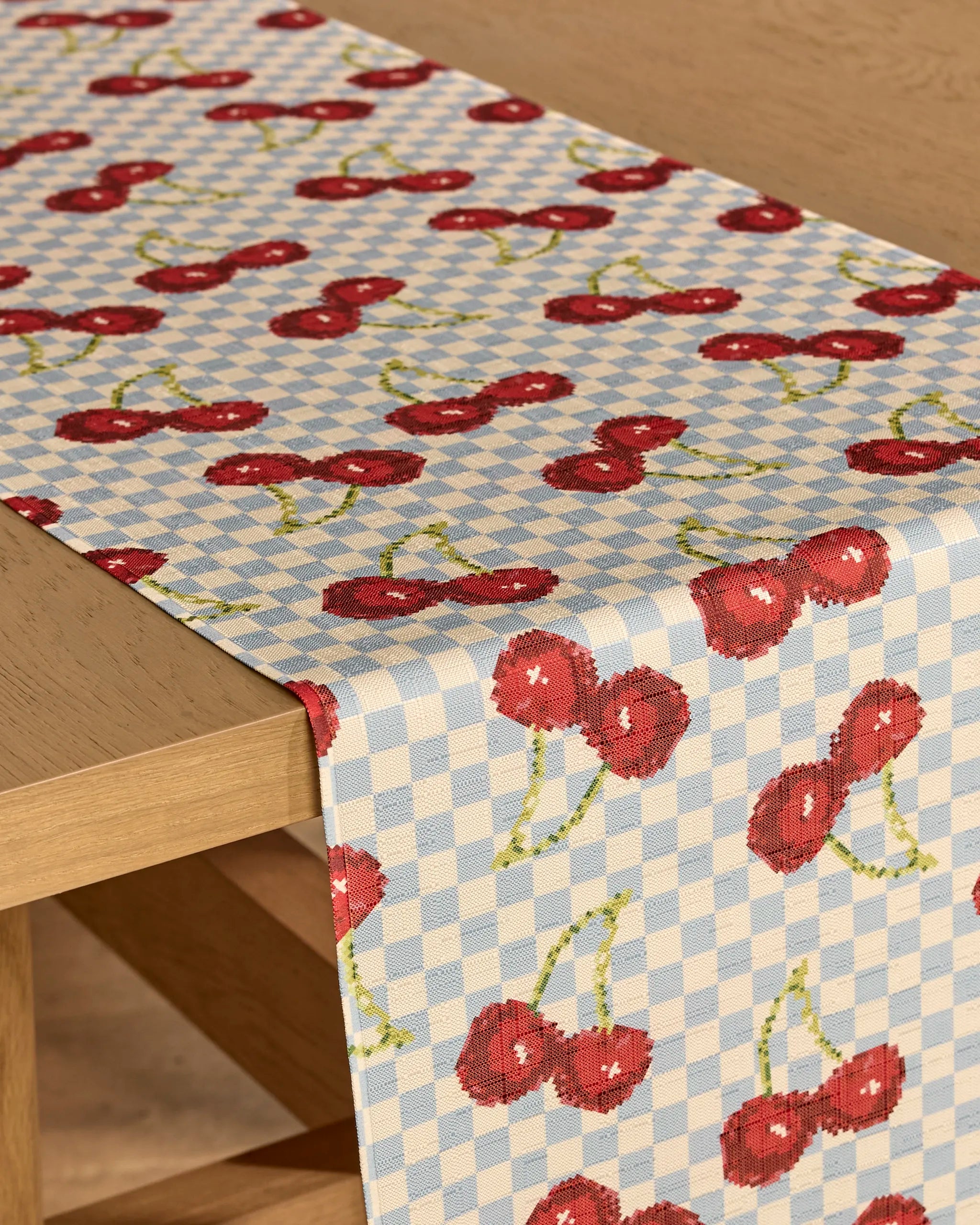 The Geometry "Cherry Cola Days" table runner features a stain-resistant, light blue and white checkered design with red cherries and green stems, elegantly draped over a wooden table.