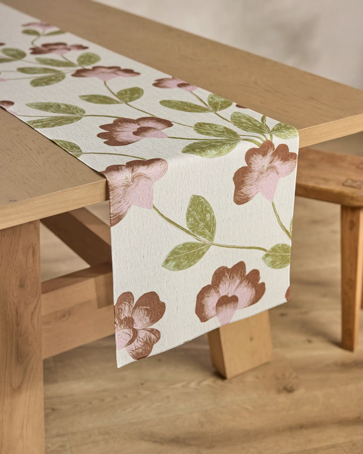 The Flower Tower table runner by Geometry, with a pink floral and green leaf botanical design, decorates a wooden table and matching bench. The stain-resistant runner adds elegance and charm to the dining set.