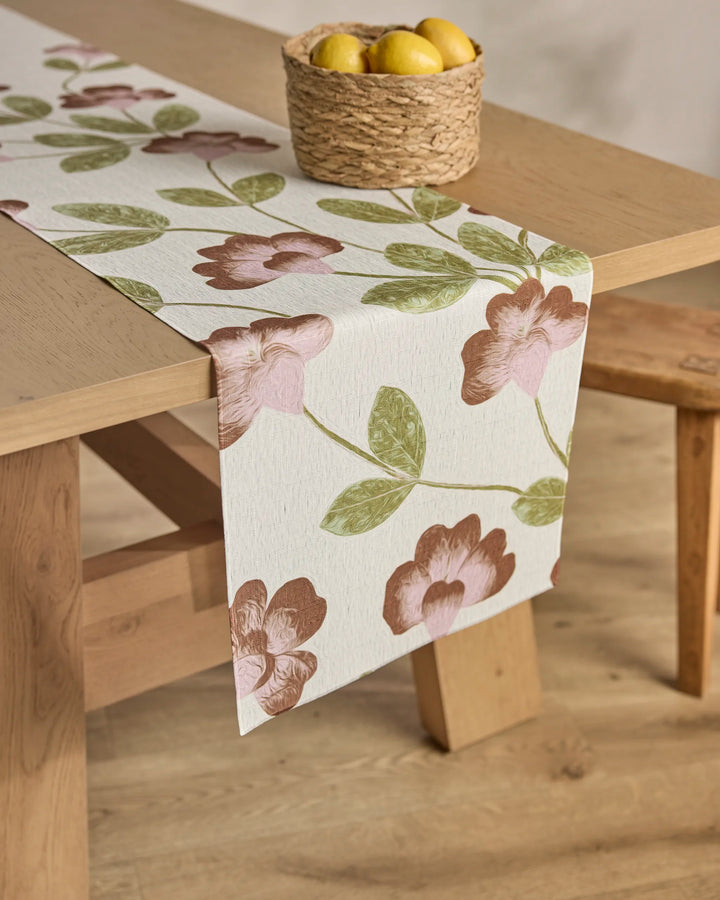 A wooden table features Geometry's Flower Tower stain-resistant table runner with pink flowers and green leaves. A woven basket of yellow lemons sits on top, and a wooden bench is placed beside the table.