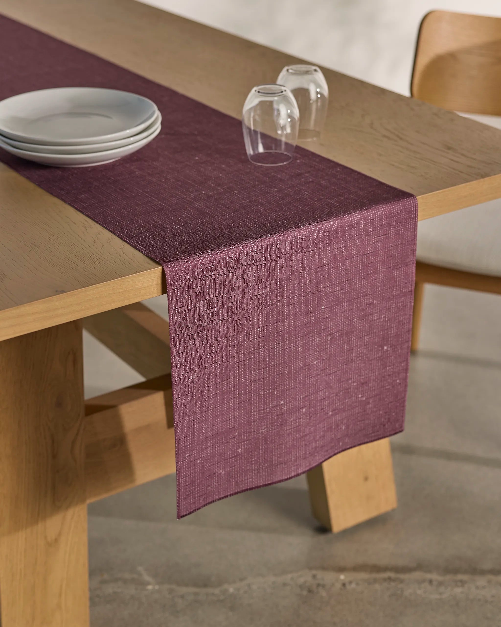 The Gather wooden dining table by Geometry features a plum stain-resistant table runner, stacked white plates, and two upside-down glasses on top, with a matching wooden chair partially visible beside the table.