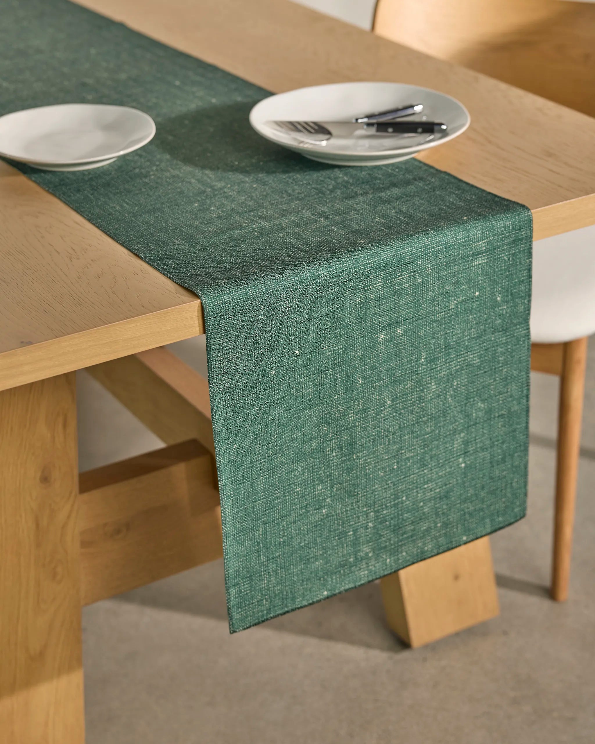 The Geometry Gather stain-resistant table runner in green rests on a light wooden dining table, set with two white plates, forks, and knives. A white chair is partially visible next to the table.