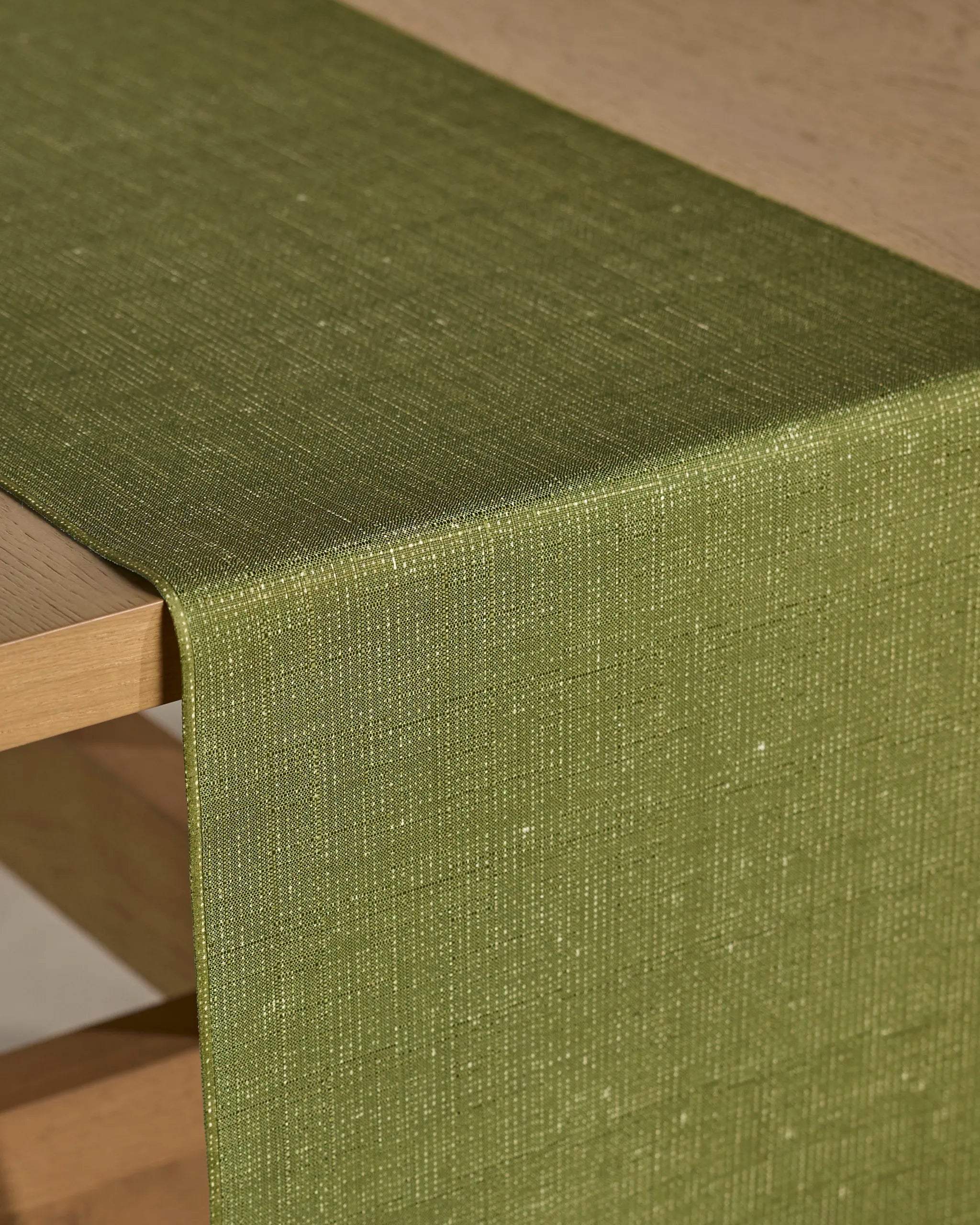 A close-up of a light wooden table featuring Geometry’s Gather table runner in textured olive green. The stain-resistant fabric has a fine woven pattern and a subtle sheen, draped elegantly over the edge.