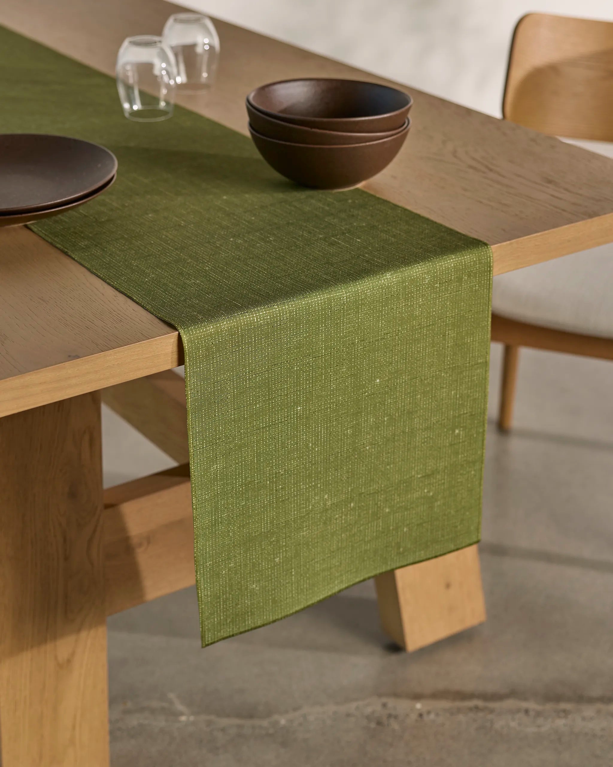The Geometry Gather dining set features a wooden table with a green stain-resistant runner, two stacked brown bowls, a brown plate, two upside-down glasses, and a matching wooden chair on a light-colored floor.