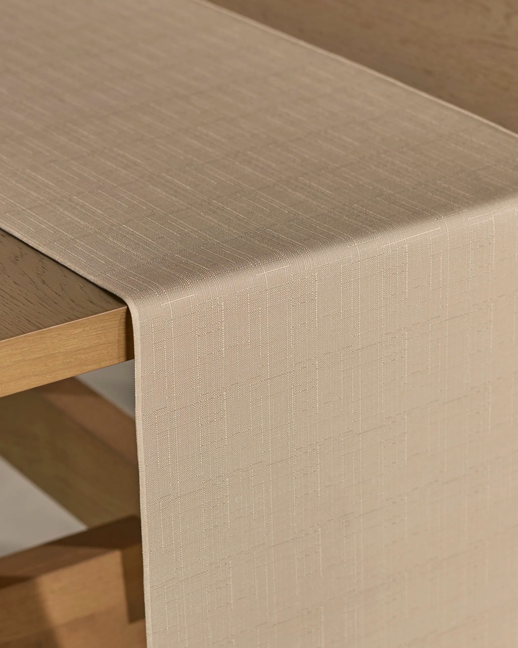 The Geometry Gather table runner, featuring a beige stain-resistant fabric with a subtle textured pattern, is draped over the edge of a light wooden table.