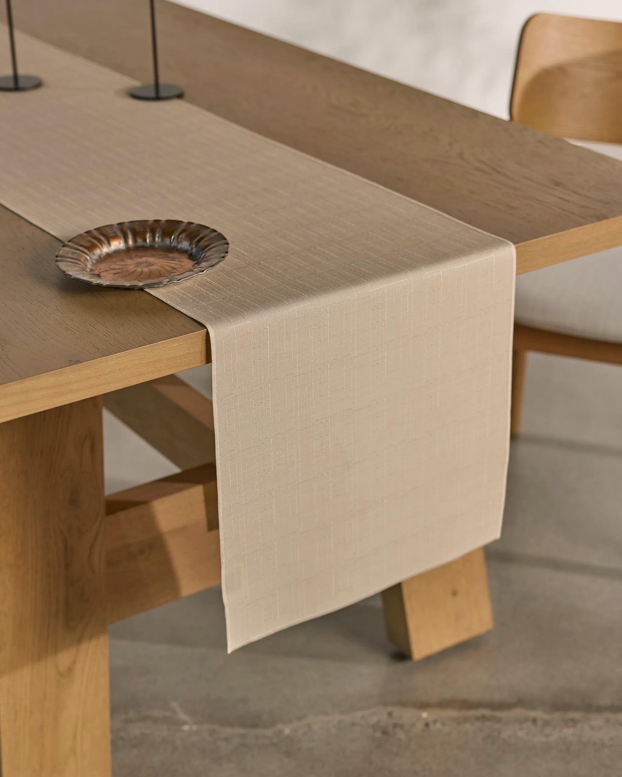 A stain-resistant, light beige Geometry Gather table runner is draped over a wooden dining table with a matching chair. A decorative metal plate rests on the runner, creating a minimal, modern look.