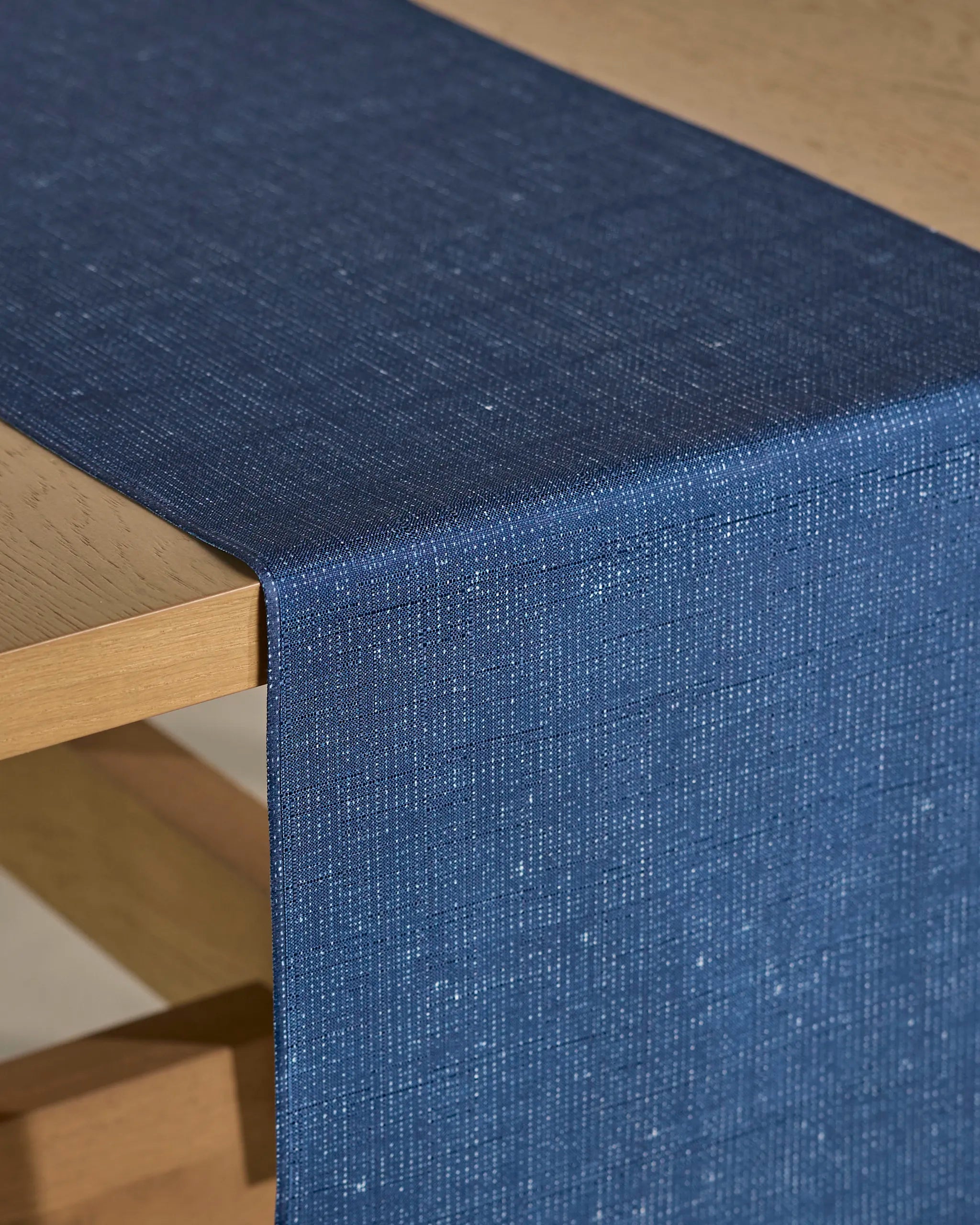 A close-up of a wooden table with Geometry's Gather table runner, featuring a textured, dark blue woven fabric made from recycled materials, draped over the edge.