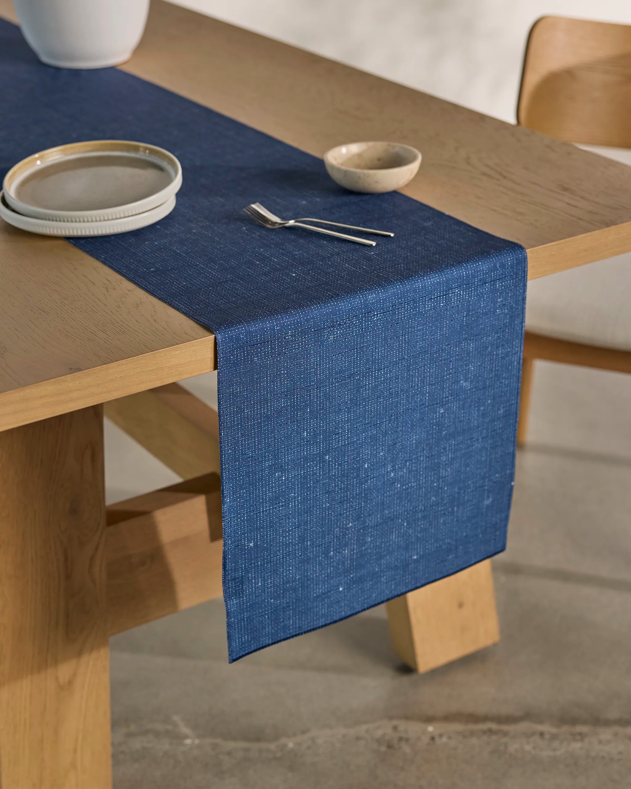 The Geometry Gather wooden dining table features a blue stain-resistant table runner, plate, fork, and small bowl. Set on a light-colored floor with a wooden chair nearby, it's perfect for stylish dining.
