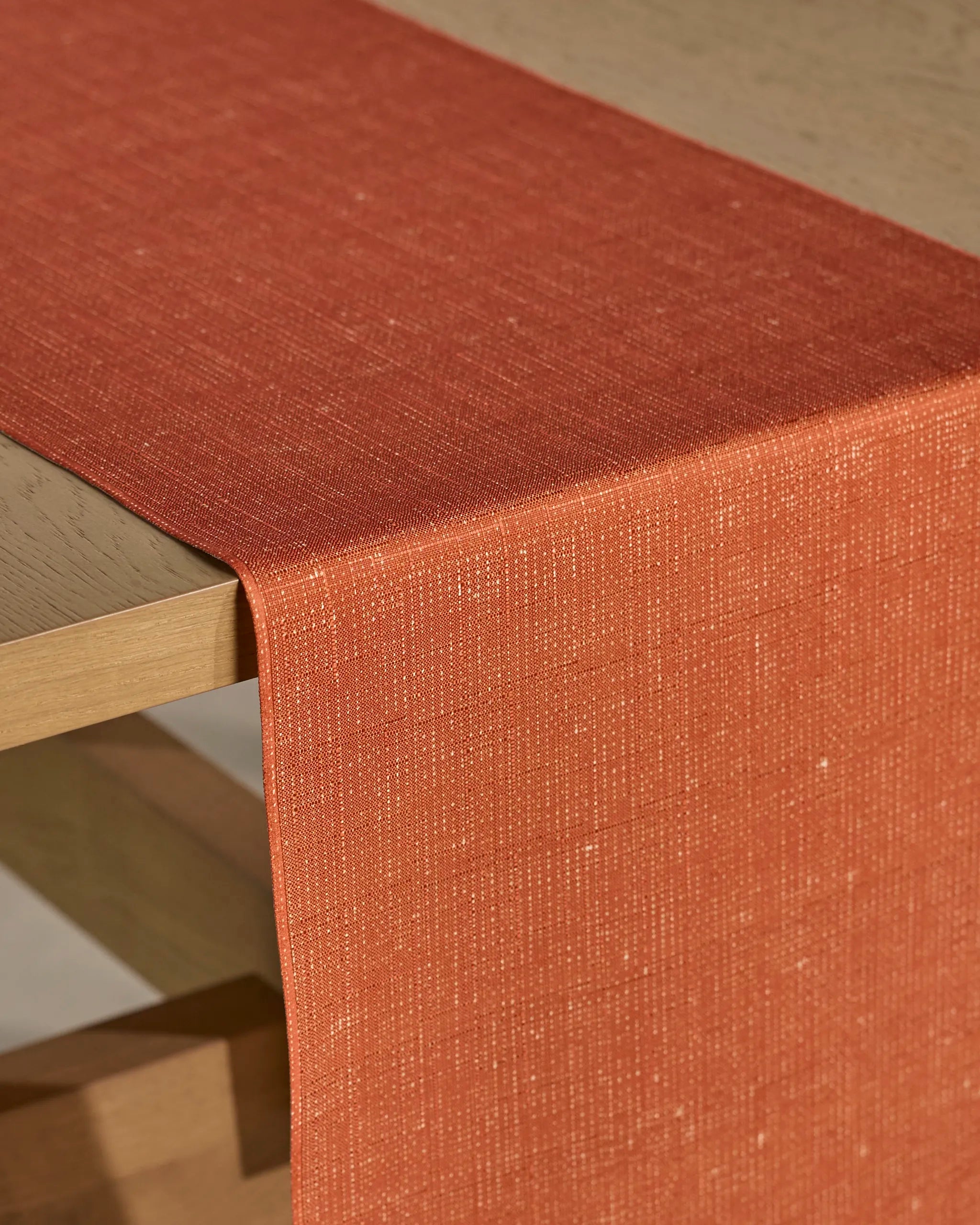 A close-up of a wooden table with the Geometry Gather table runner in vibrant orange, featuring a subtle woven pattern. This stain-resistant runner is draped neatly over the edge for added texture and modern style.