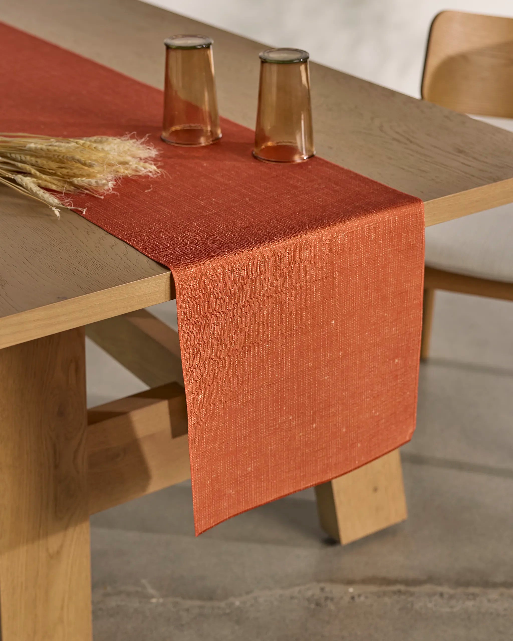 The minimal, warm-toned setting features Geometry’s Gather collection: a wooden table with an orange, stain-resistant table runner, two upside-down brown glasses, dried wheat, and a matching chair partially visible.