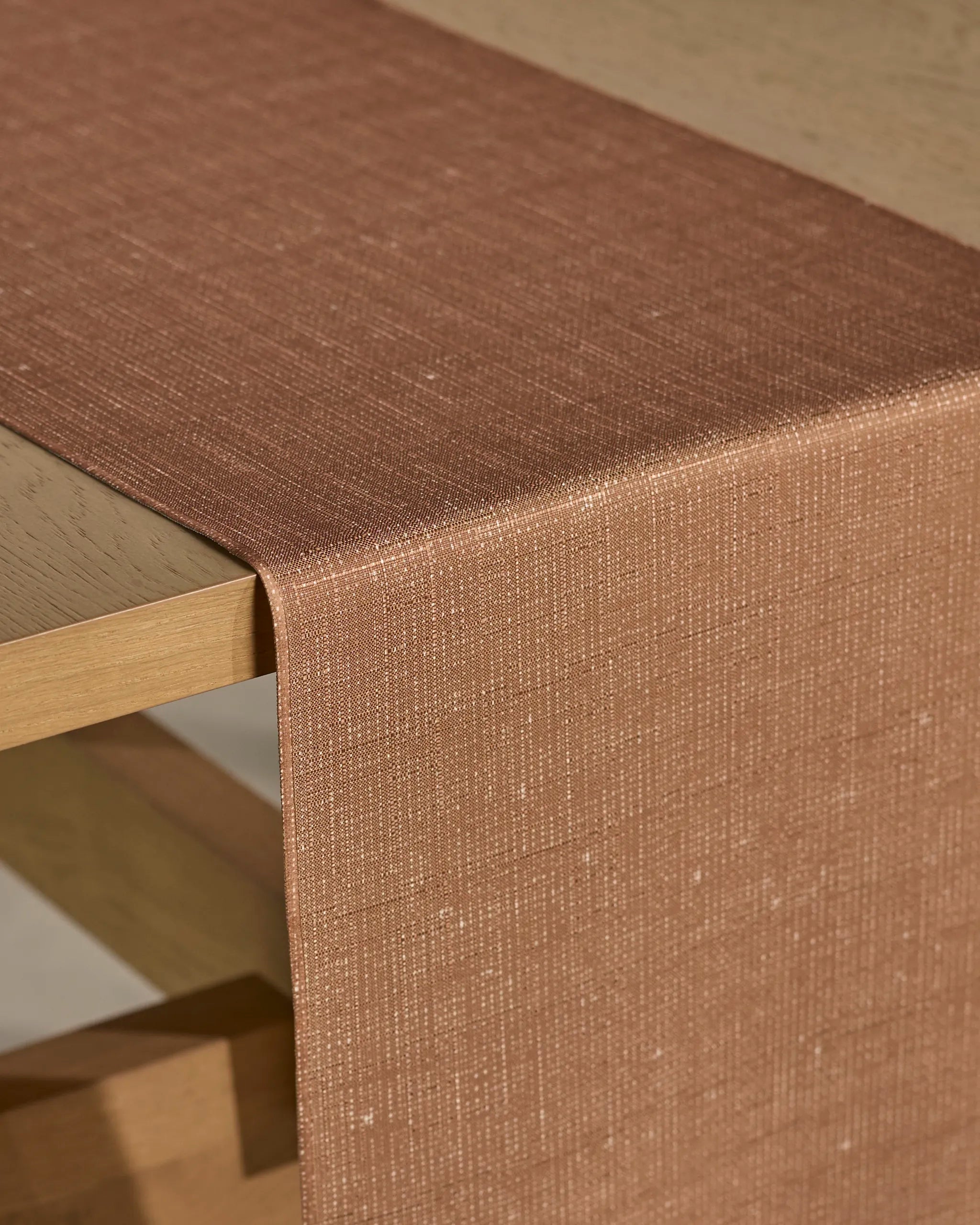 A close-up of Geometry's Gather table runner in light brown, draped over a light wooden table, highlights its textured, stain-resistant fabric.
