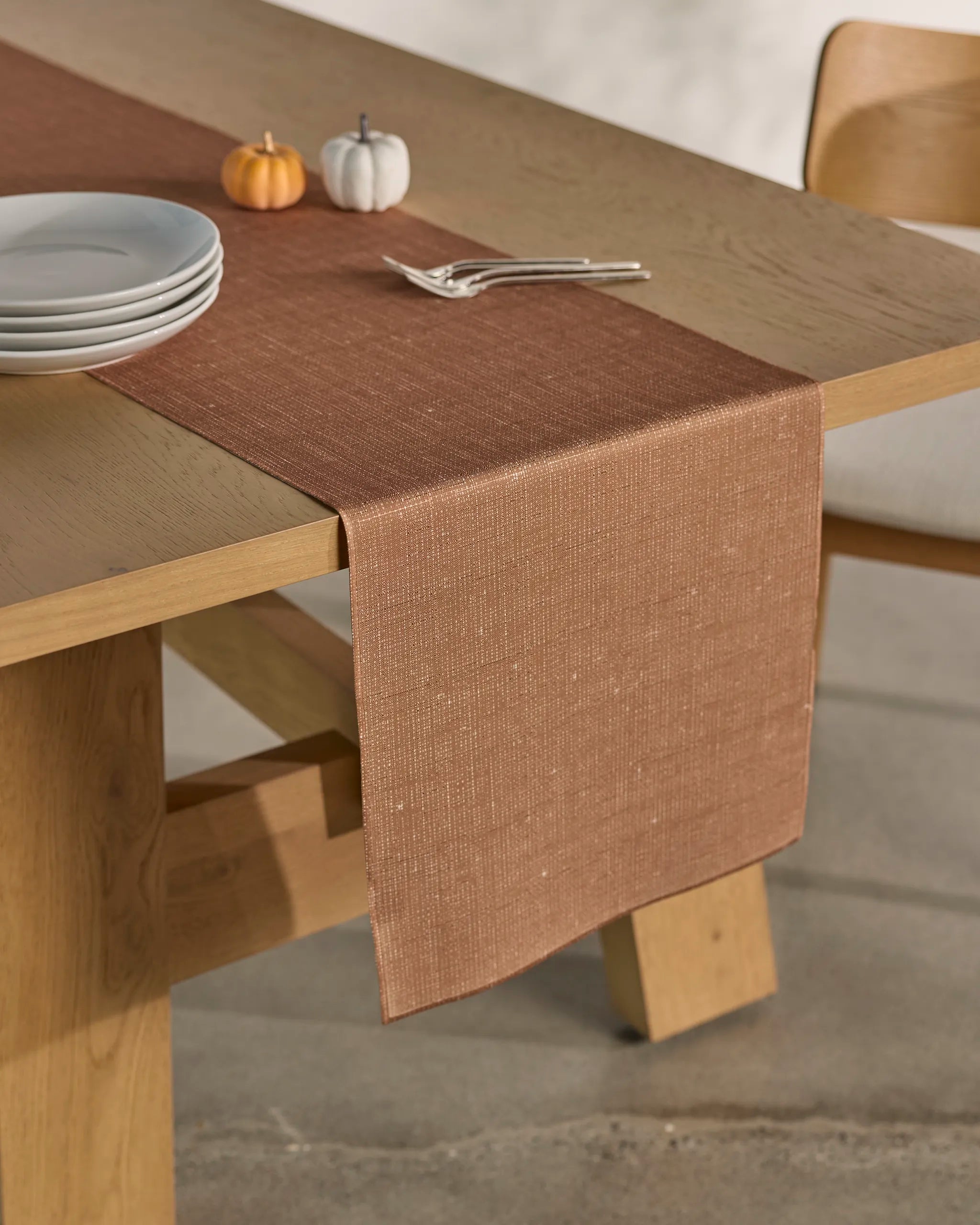 The Geometry Gather light wooden dining table features a stain-resistant runner, stacked white plates, two small decorative pumpkins (one orange, one white), and two neatly arranged forks.