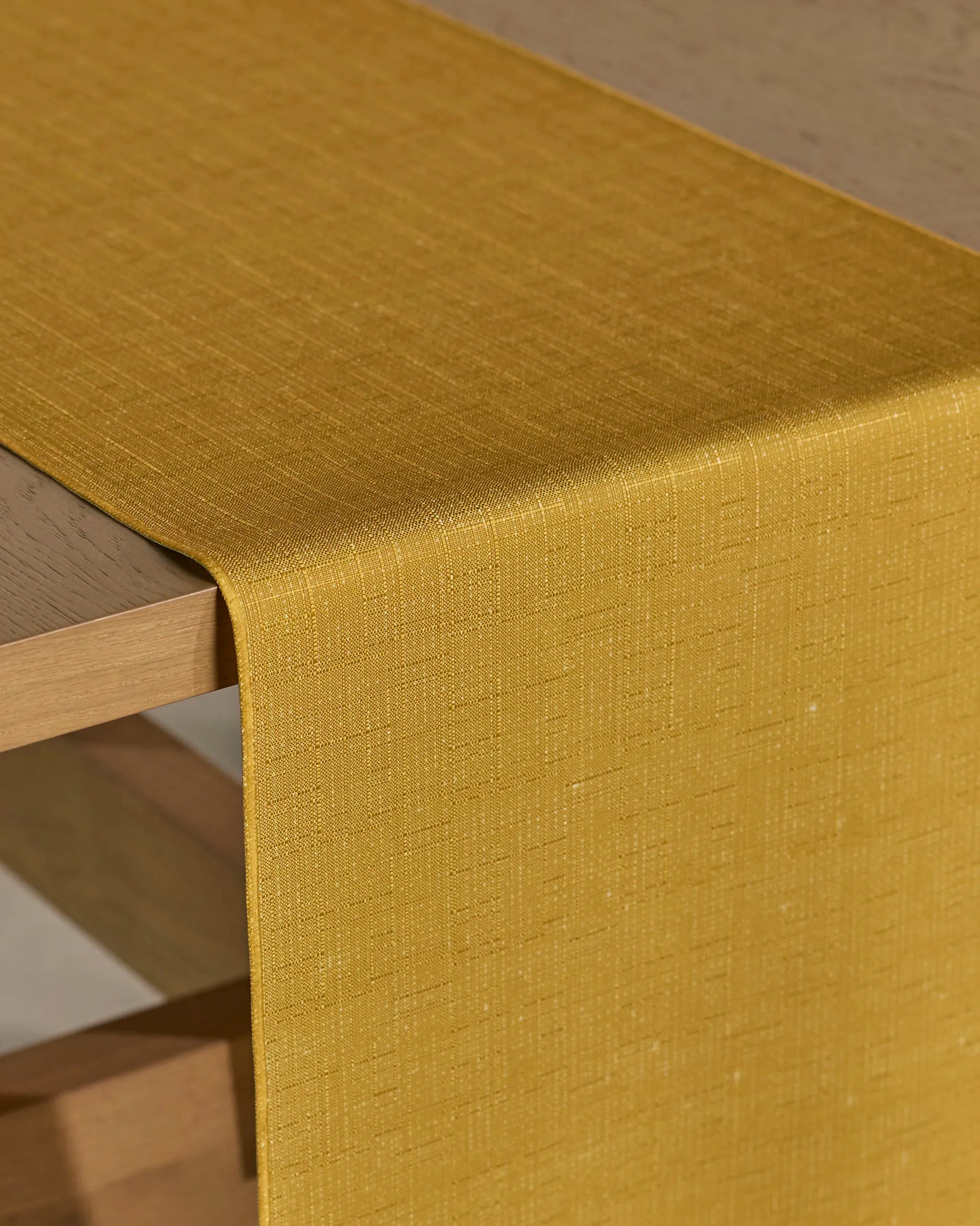 The Geometry Gather table runner, in stain-resistant mustard yellow with a subtle textured pattern, is neatly draped over the corner of a light wooden table.
