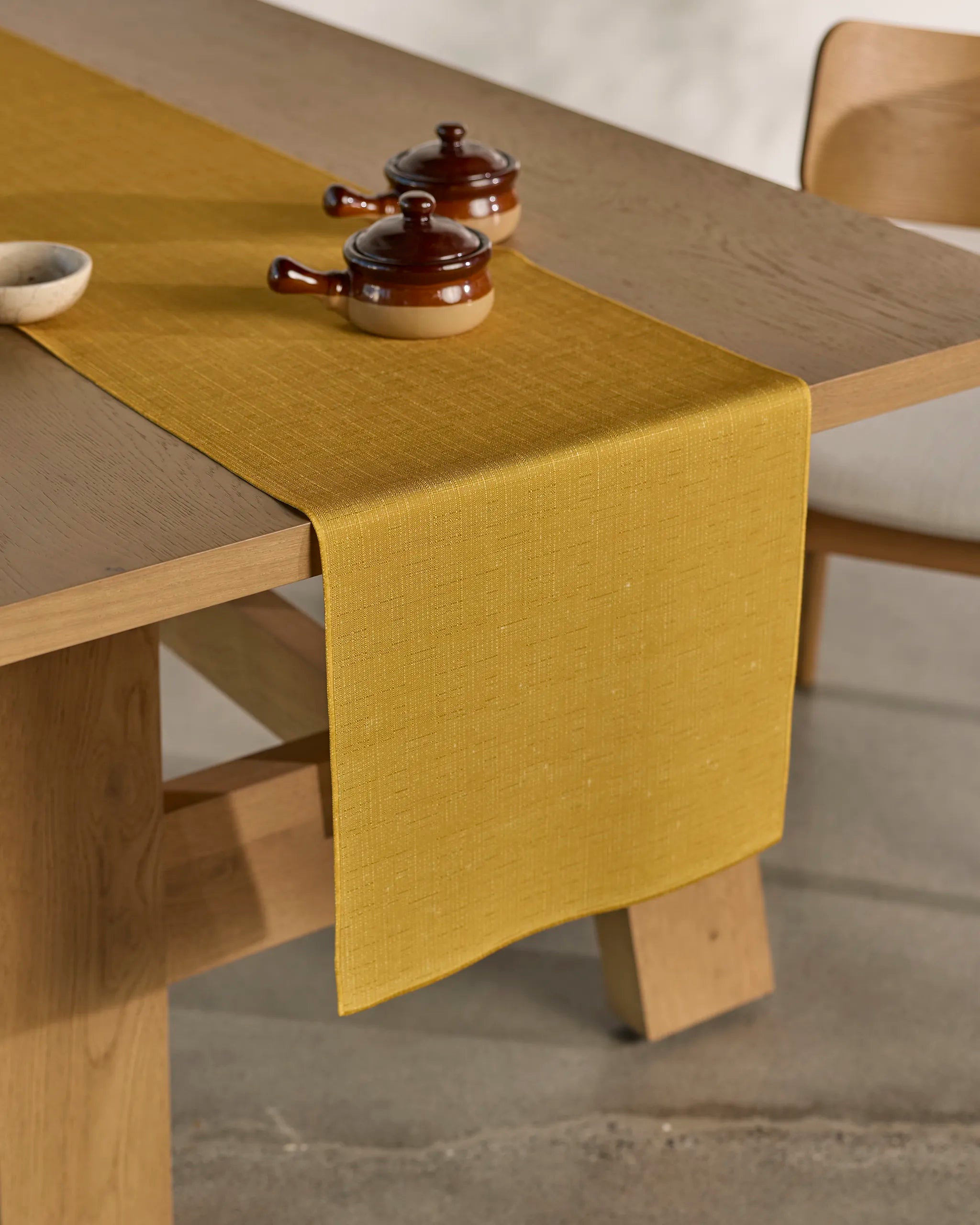 The Geometry Gather wooden dining table features a stain-resistant mustard yellow table runner, two small lidded brown ceramic dishes, and part of a wooden chair nearby.