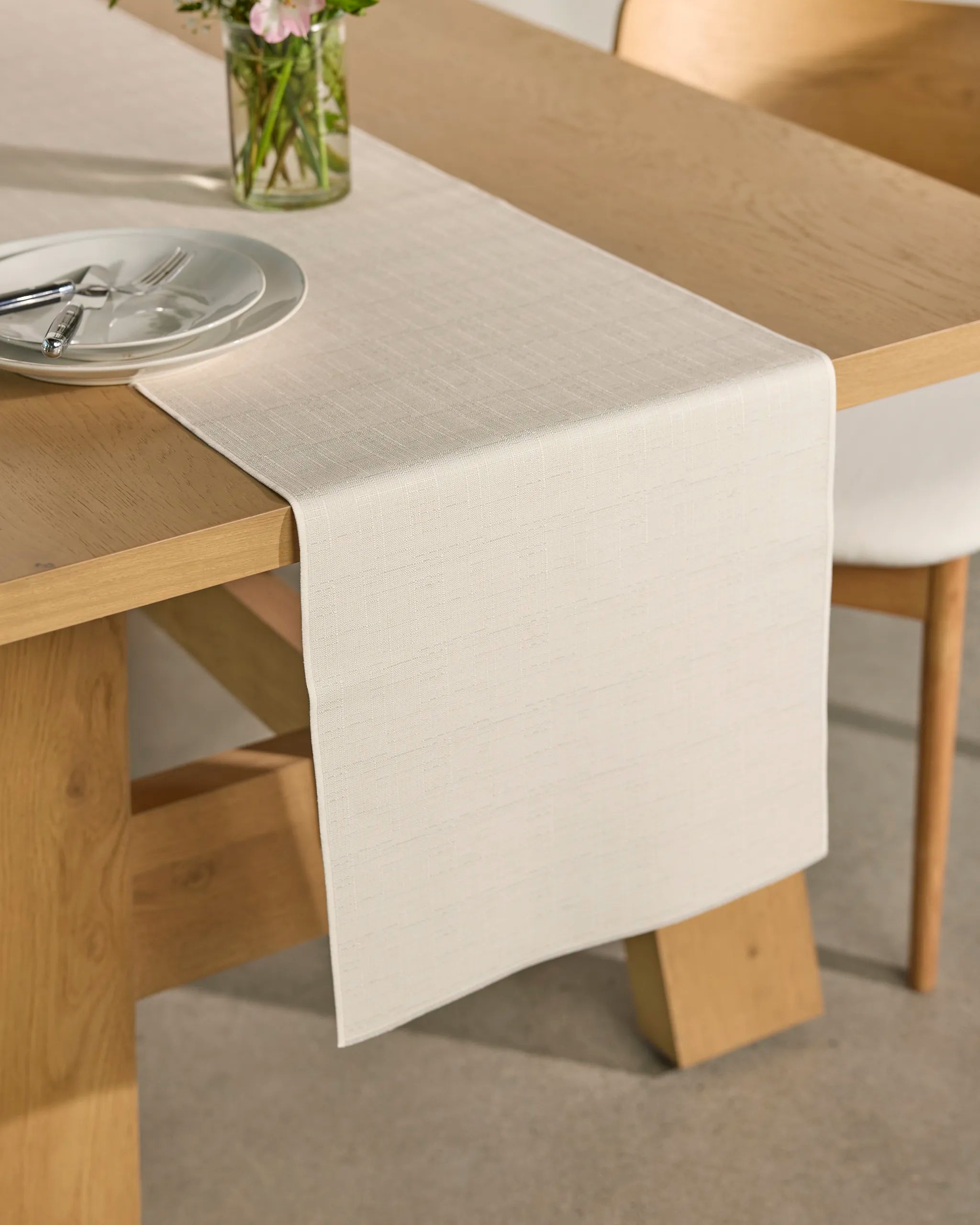 A light beige “Gather” table runner by Geometry, known for its stain-resistant fabric, drapes over a wooden dining table set with a white plate and silverware. In the background, a glass vase holding pink and white flowers completes the scene.