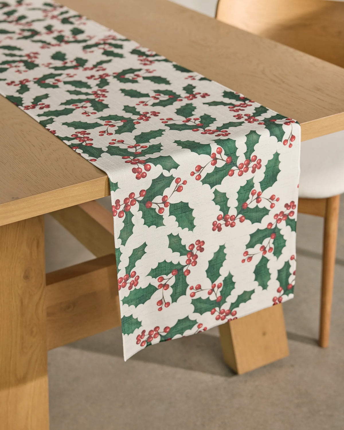 A wooden table features the Geometry “Holly Days” white, stain-resistant table runner adorned with green holly leaves and red berries, with a light-colored chair positioned beside it.