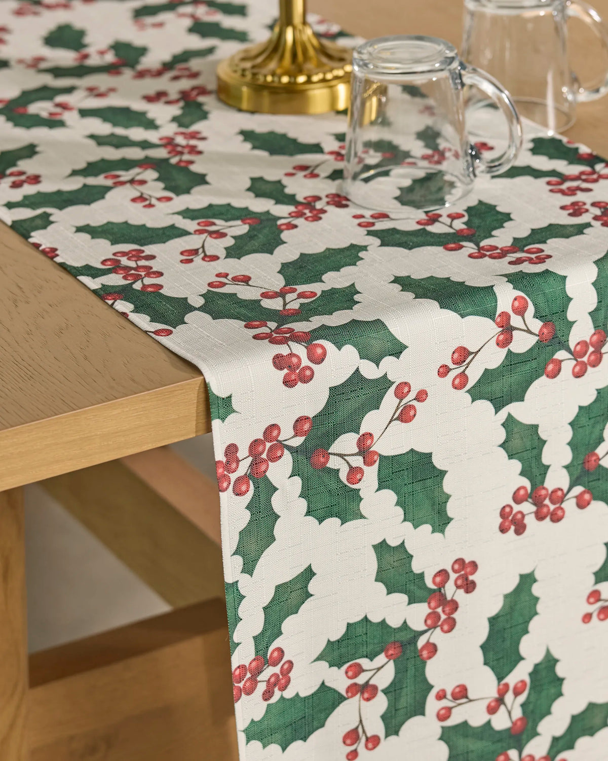 A Geometry “Holly Days” stain-resistant table runner featuring a holly leaf and red berry pattern is displayed on a wooden table, accompanied by glass mugs and a brass candlestick.