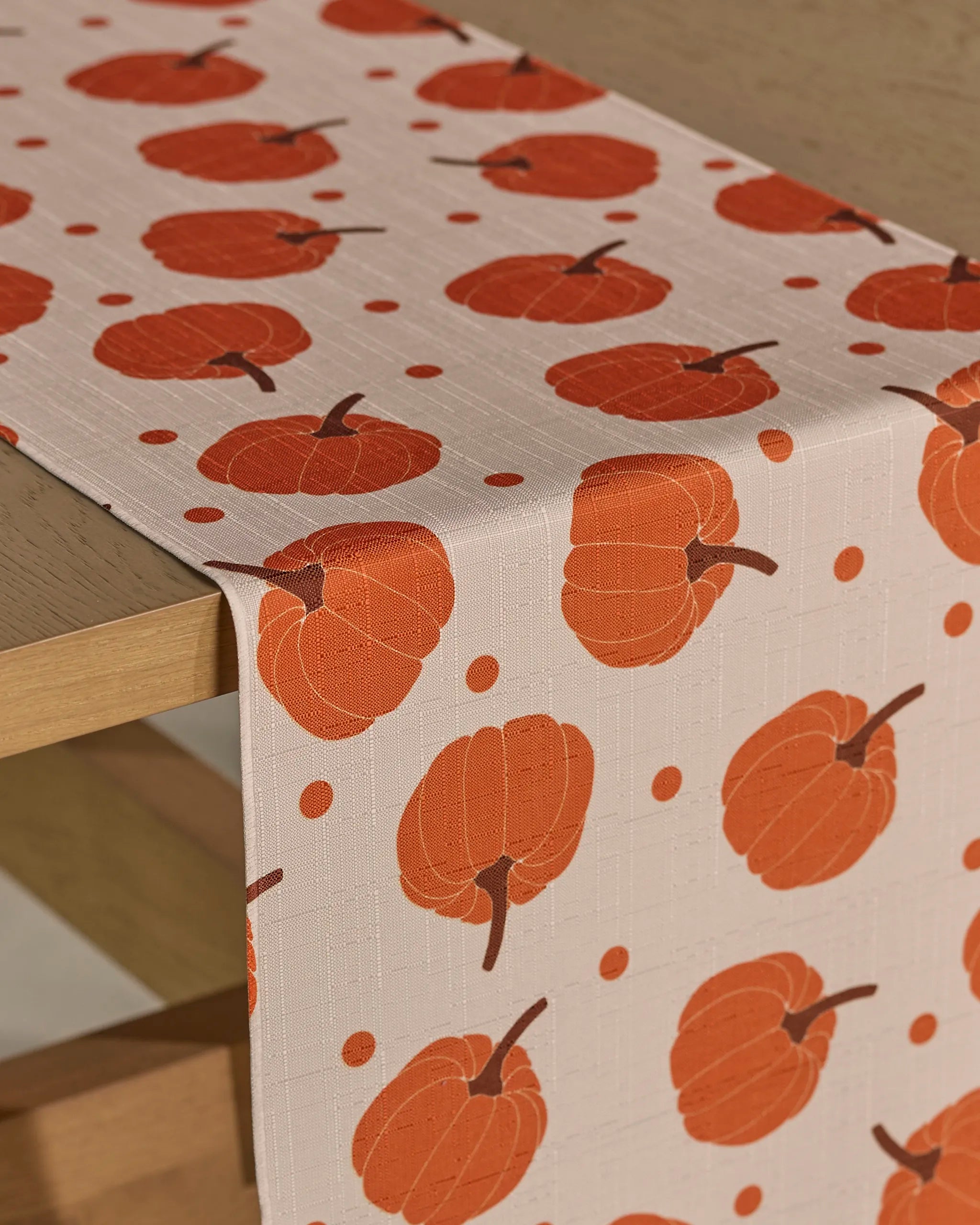 The Geometry Pumpkin Polka table runner, in beige with an orange pumpkin and dot pattern, offers stain resistance and is draped over a wooden table.