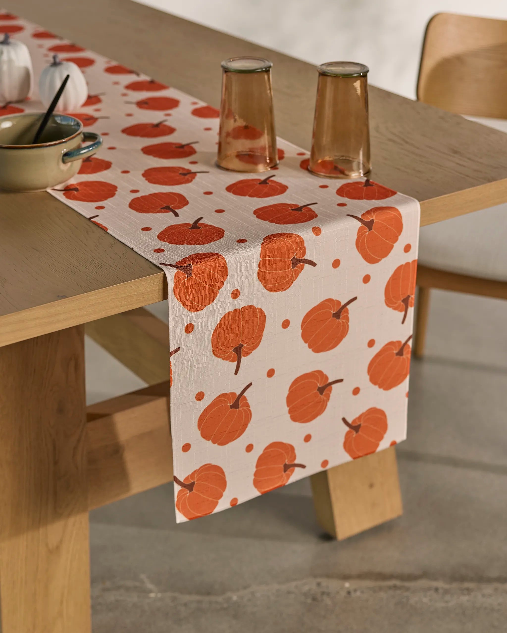 A wooden table with a Geometry Pumpkin Polka stain-resistant table runner featuring a white and orange pumpkin pattern, two glasses of light drink, a bowl with a spoon, and two small white pumpkin decorations.