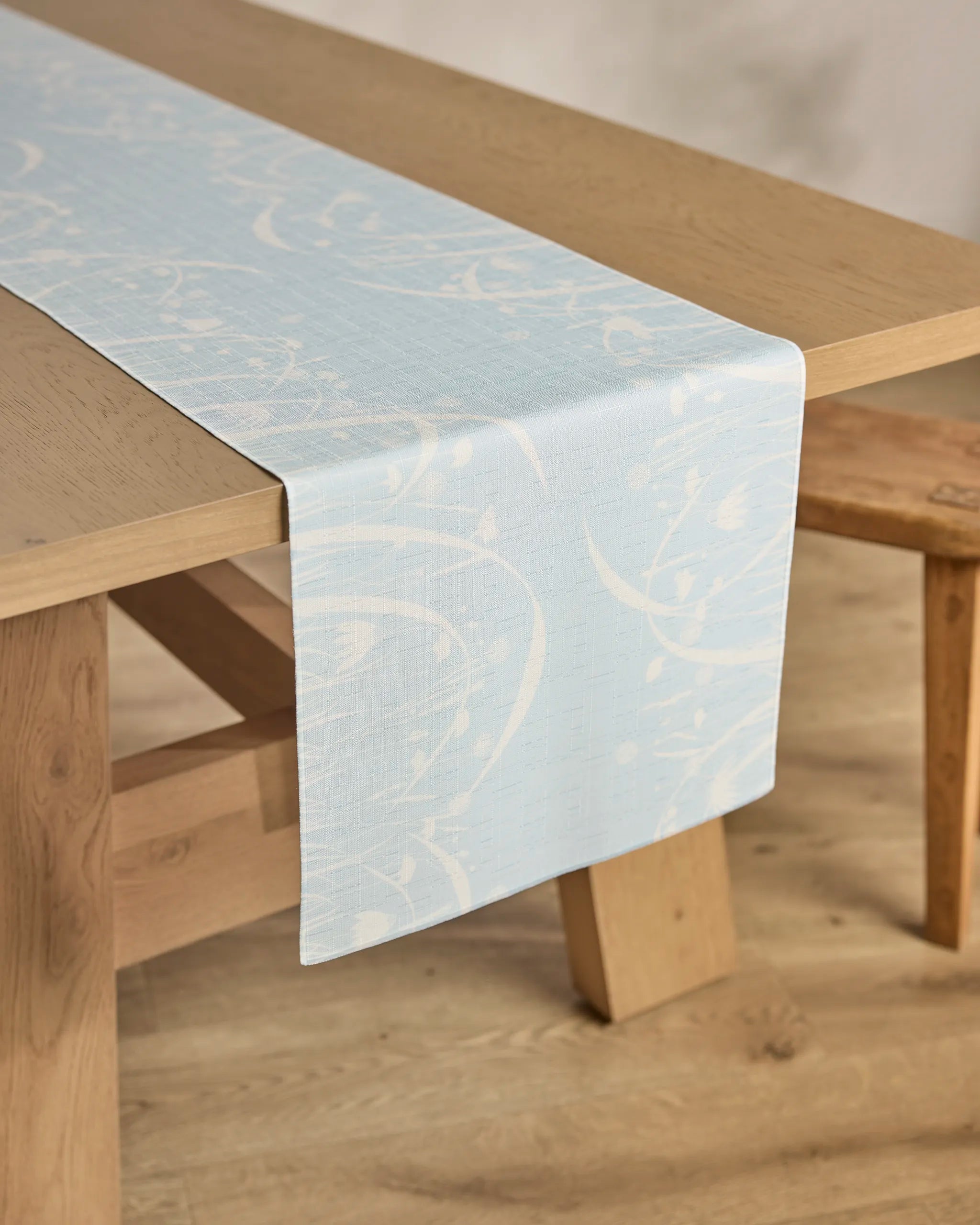 The Geometry Quiet Bloom table runner, featuring a light blue stain-resistant fabric with a subtle white floral pattern, is draped over a wooden dining table and bench. Its practical elegance stands out against the neutral background.