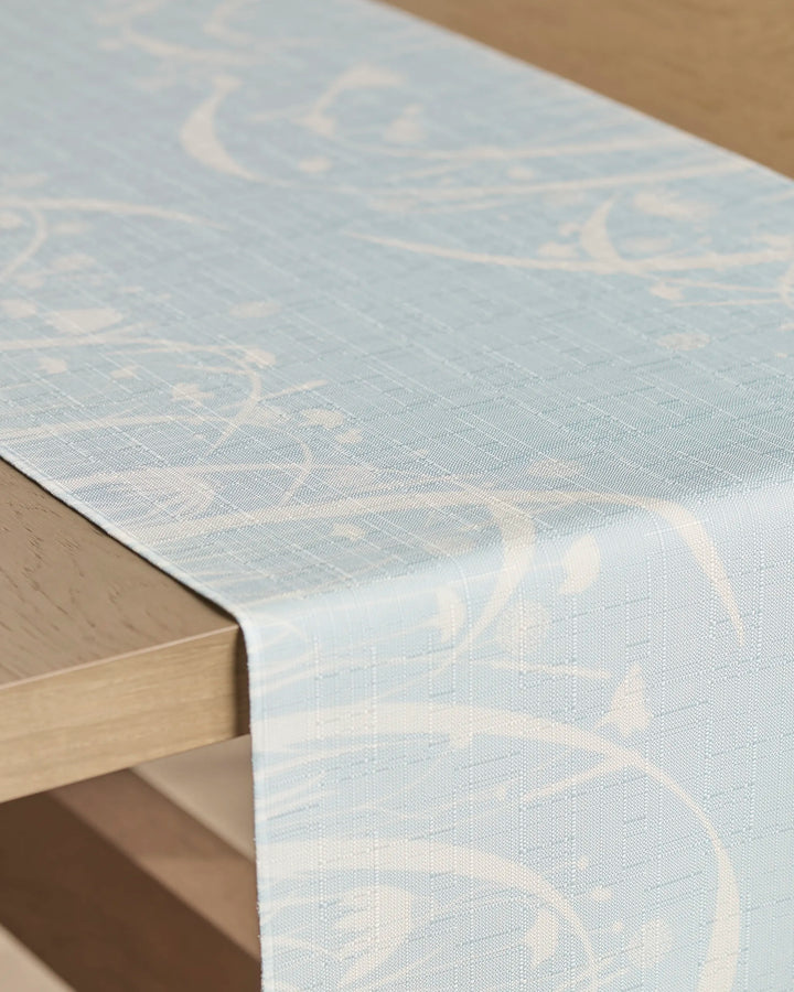 A close-up of the Geometry Quiet Bloom table runner, featuring a light blue and white swirling pattern. Stain-resistant, it’s draped over a light wood table.