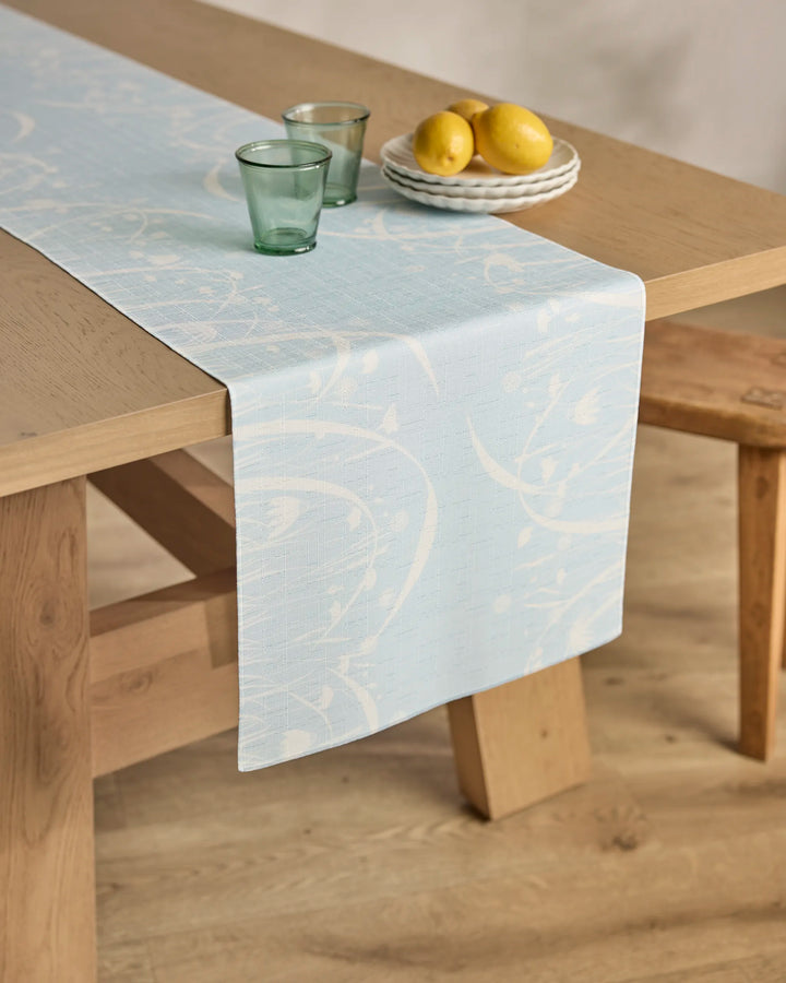 A wooden table features the Quiet Bloom table runner by Geometry in a light blue pattern, two green glass tumblers, and white plates with three lemons. A wooden bench stands next to the table on a light wood floor.