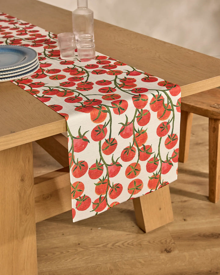 A wooden dining table with the Geometry "Ripe and Ready" stain-resistant table runner, featuring a vibrant tomato pattern, set with stacked blue plates, clear glasses, and a glass bottle.