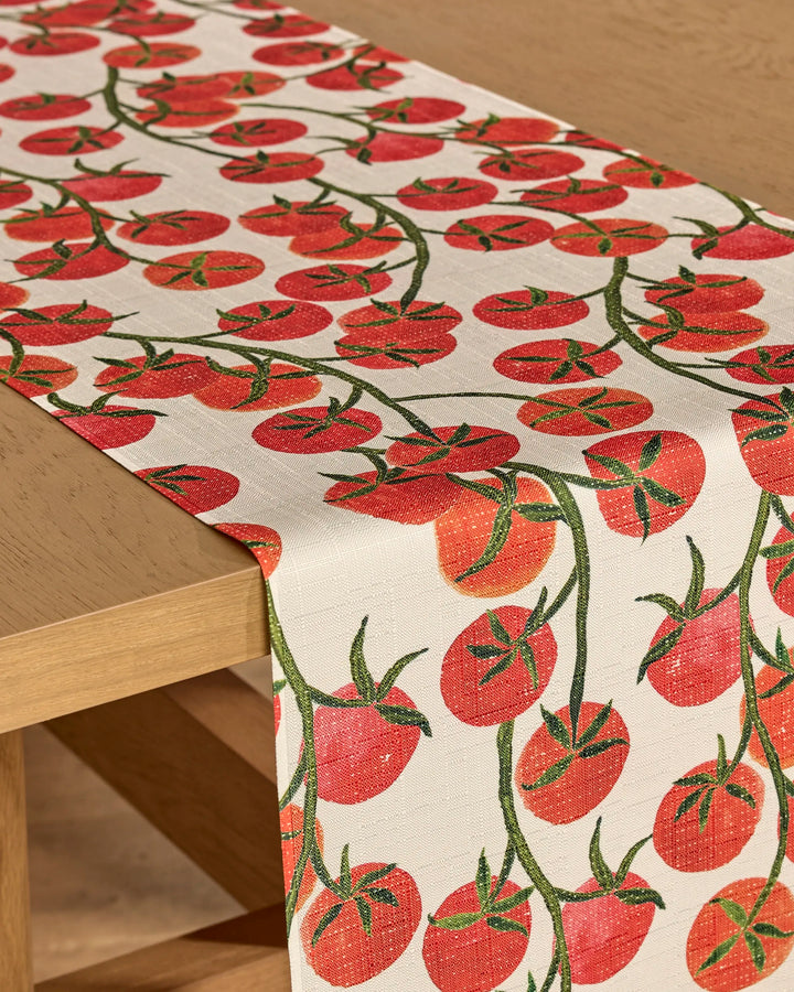 The Ripe and Ready table runner by Geometry, featuring a colorful tomato vine pattern with red tomatoes and green stems, adds a lively touch to your light wooden table while offering beige, stain-resistant durability.