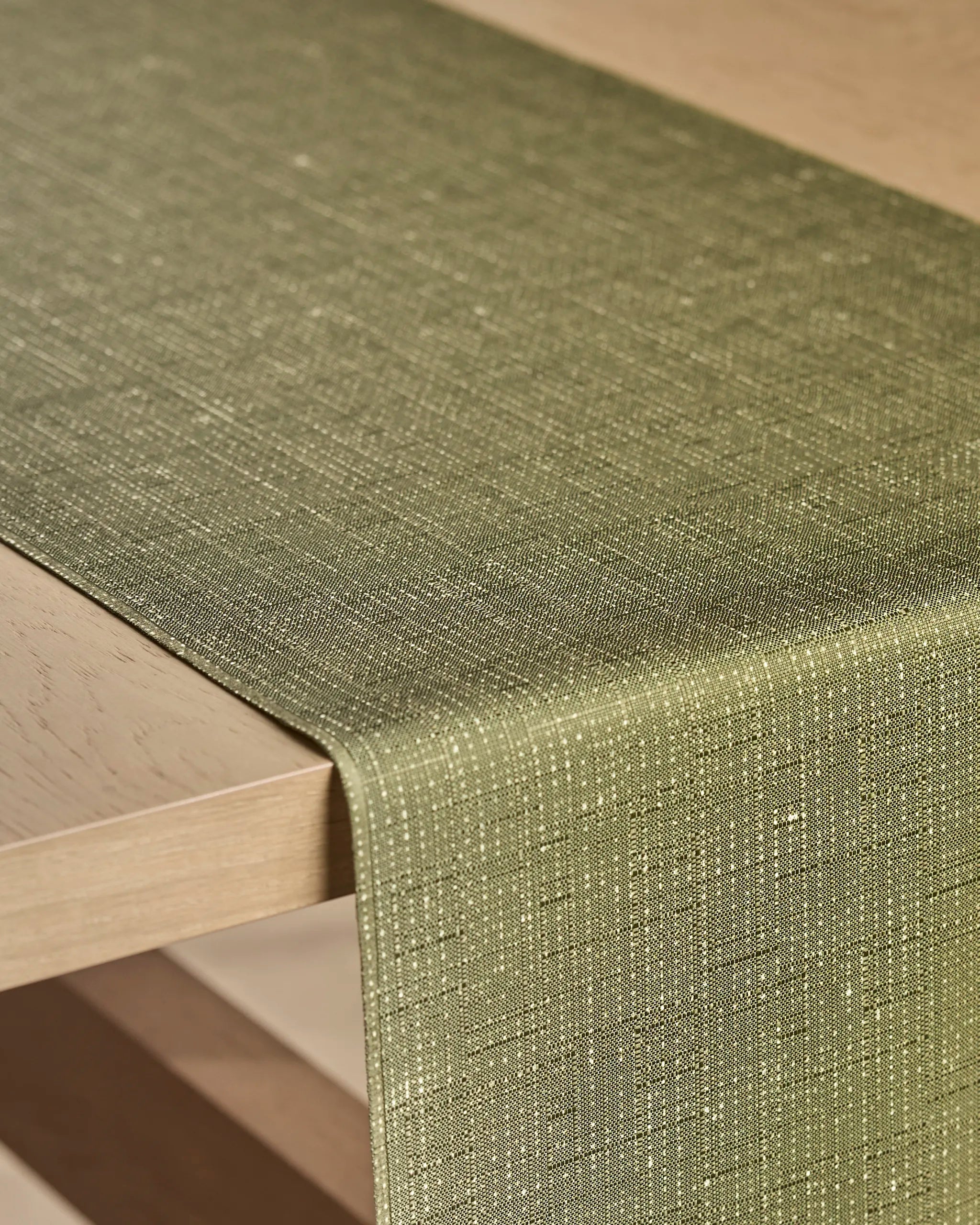 A close-up of the Willa Green recycled table runner by Geometry, featuring a textured green fabric with subtle metallic threads, draped over the edge of a light wooden table.