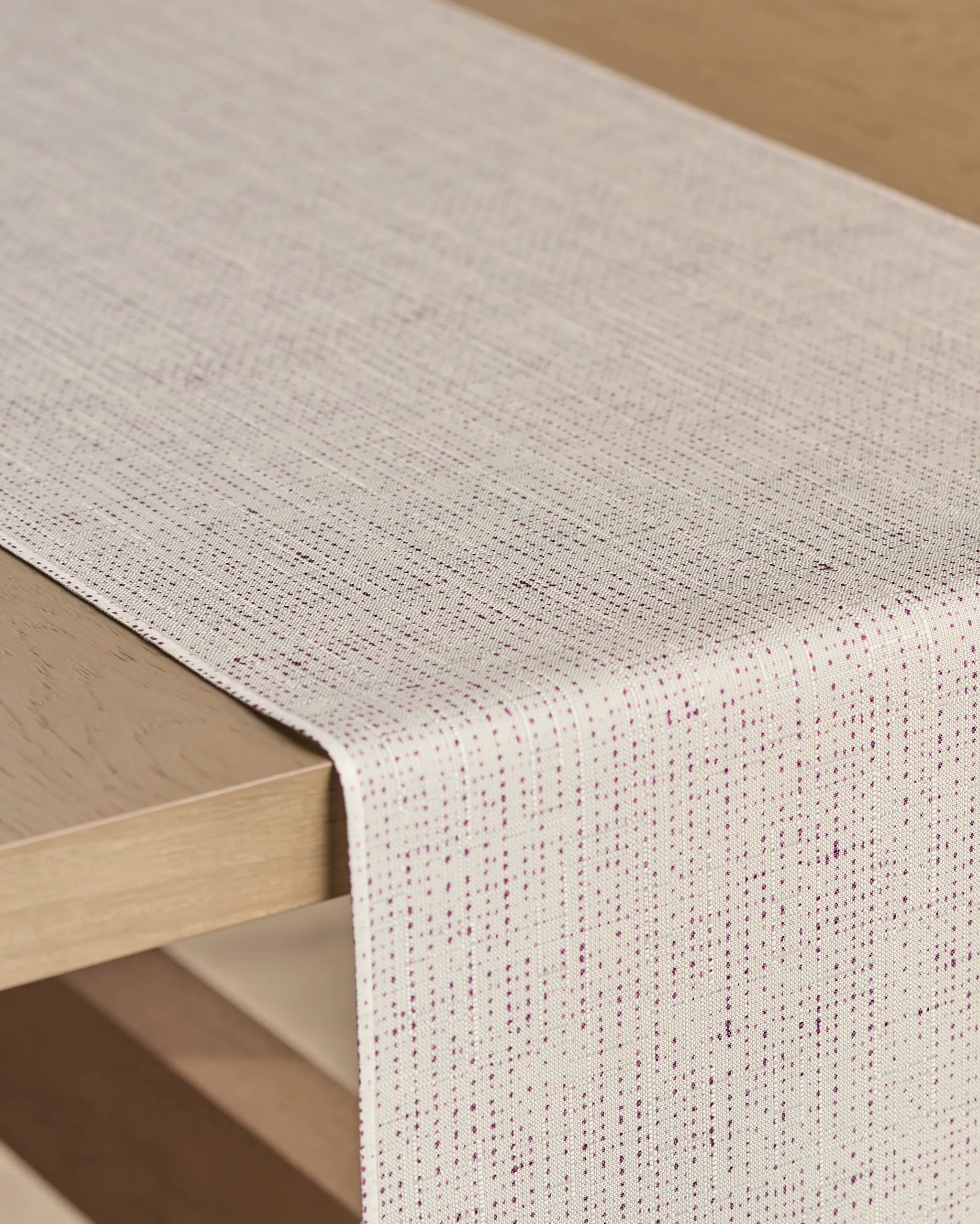 A close-up of the Geometry Willa Lav stain-resistant table runner, featuring a subtle speckled pattern, draped over the edge of a light wooden table.