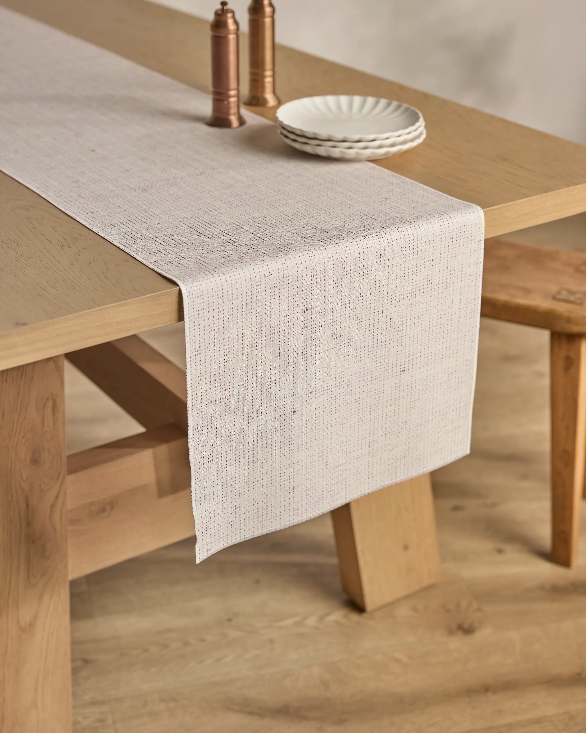 The Willa Lav table runner by Geometry, in light beige and stain-resistant fabric, drapes over a wooden dining table set with white plates and copper salt and pepper mills. A wooden bench sits beside the table on a light wood floor.